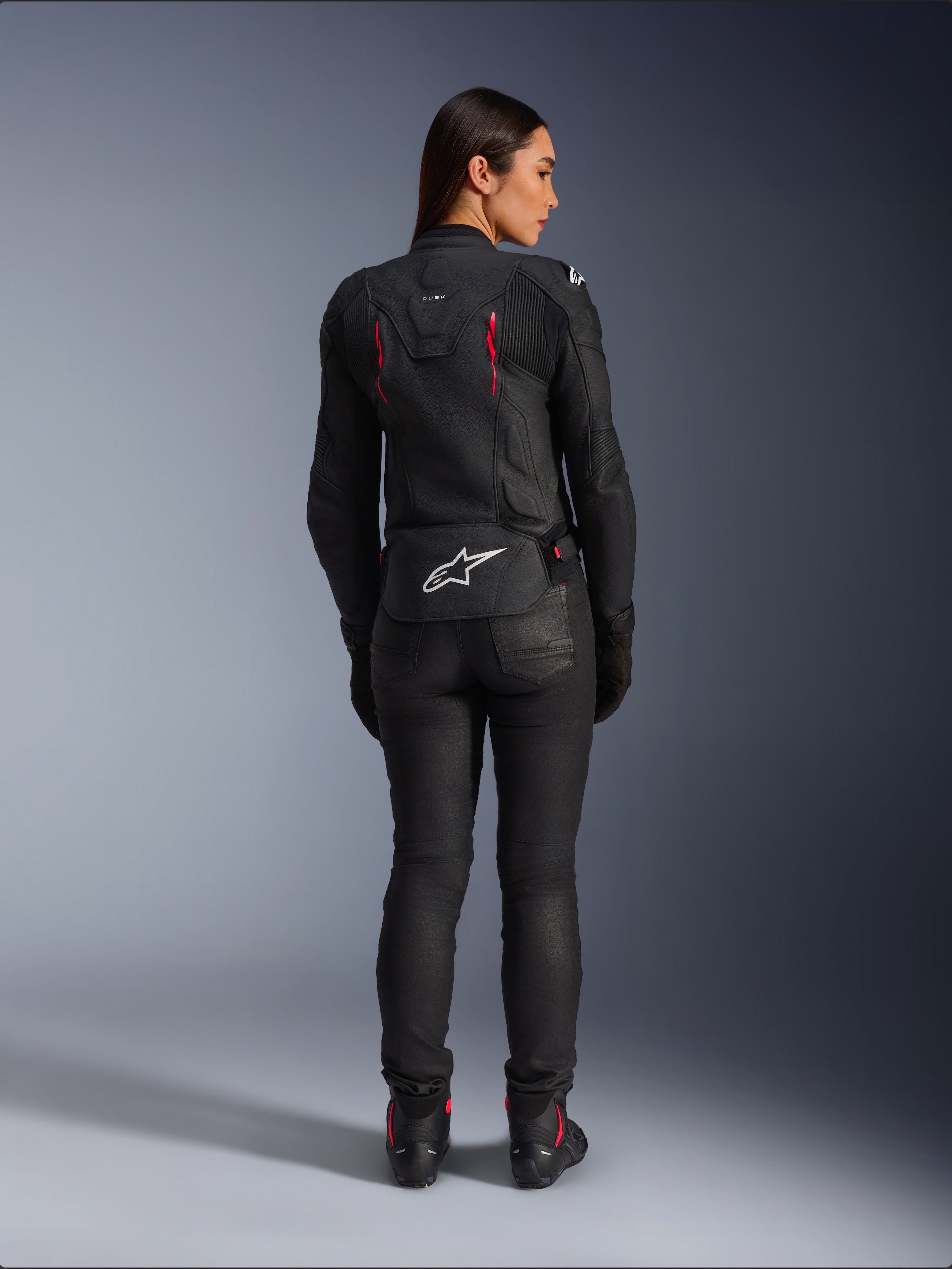 Alpinestars Stella Dusk Lederjacke, Rückansicht, schwarzes Leder mit Akzenten in Diva Pink und Weiß, weißes Alpinestars Logo am unteren Rücken und pinke Belüftungsdetails, getragen mit schwarzer Motorradhose und Stiefeln, hochwertige Sport-Motorradausrüstung für Damen