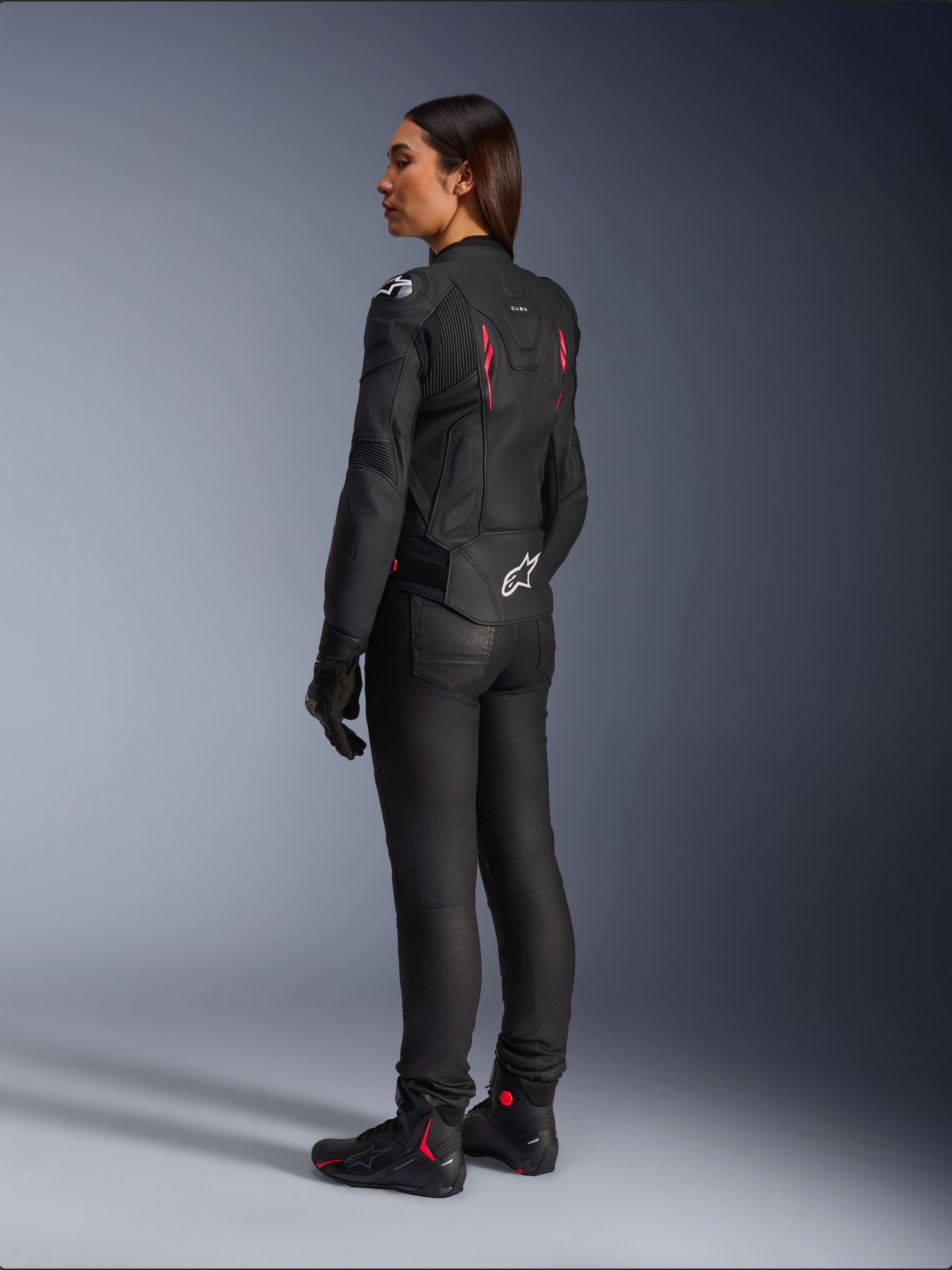 Stella Dusk Damen-Lederjacke, Schwarz Diva Pink Weiß, Dreiviertel-Rückansicht an einer Fahrerin, kombiniert mit schwarzer Motorradhose und schwarzen Stiefeln, hochwertige Lederkonstruktion mit pinken Akzenten und weißem Alpinestars-Logo am unteren Rücken