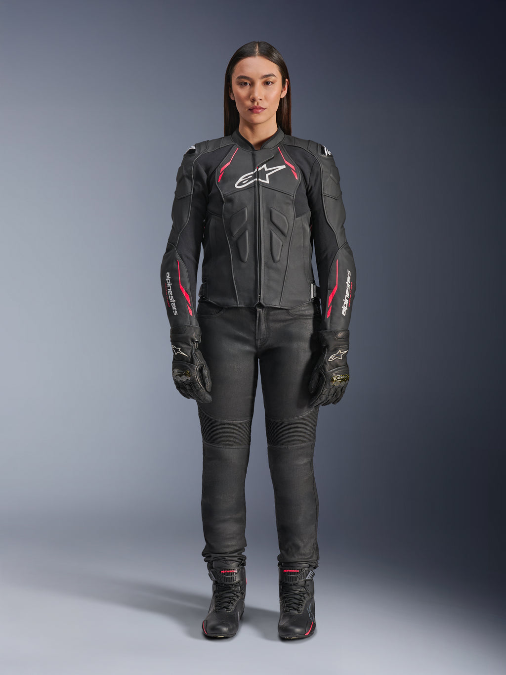 Eine Frau trägt die Alpinestars Stella Dusk Lederjacke in Black Diva Pink White, mit einem weißen Brustlogo und pinkfarbenen Akzenten an den Schultern und Ärmeln, kombiniert mit einer schwarzen Motorradhose, Handschuhen und Stiefeln.