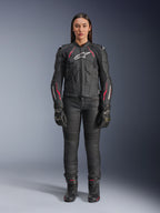 Eine Frau trägt die Alpinestars Stella Dusk Lederjacke in Black Diva Pink White, mit einem weißen Brustlogo und pinkfarbenen Akzenten an den Schultern und Ärmeln, kombiniert mit einer schwarzen Motorradhose, Handschuhen und Stiefeln.