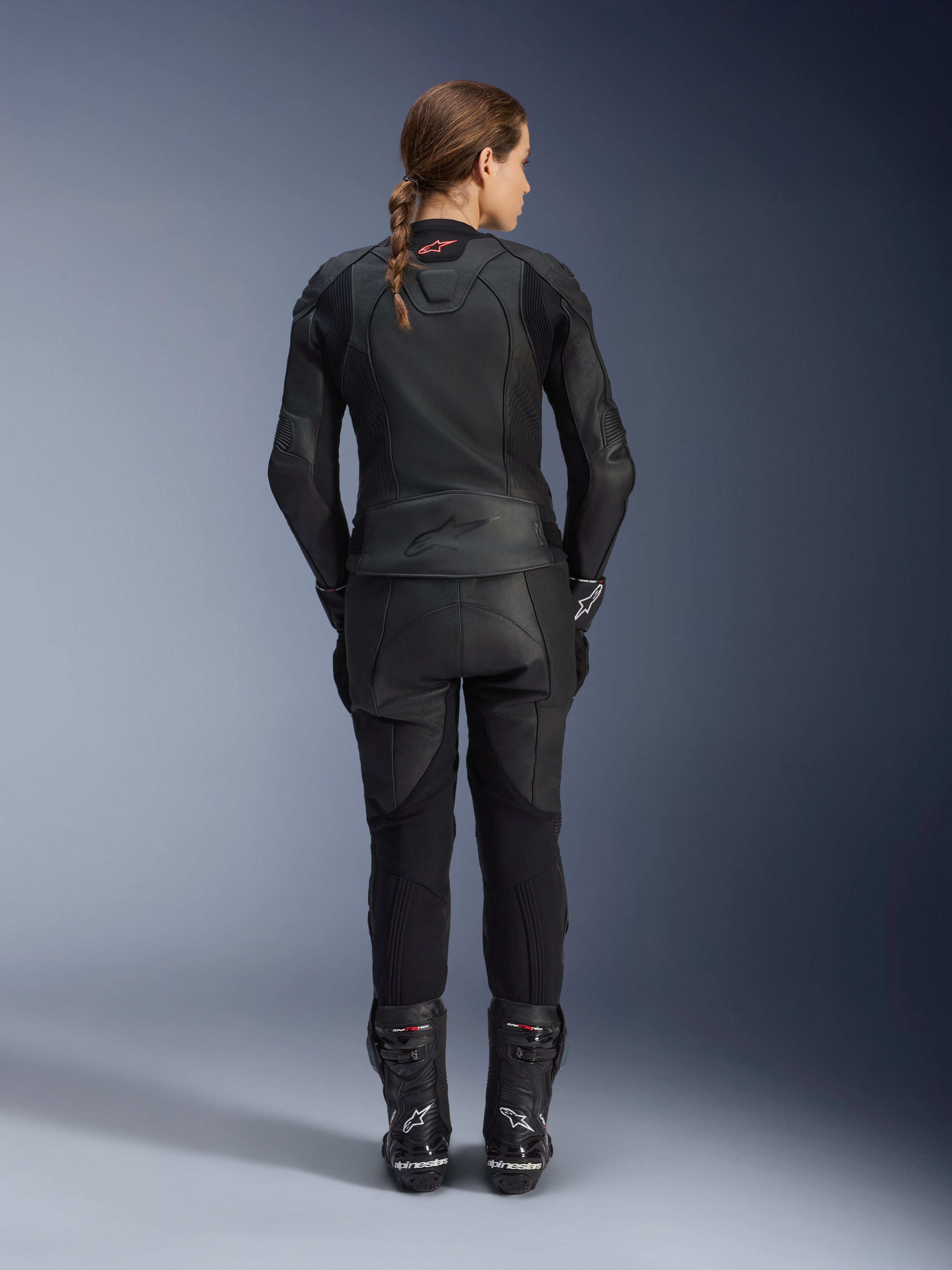 Alpinestars Stella GP Plus V4 Damen-Lederjacke, Schwarz Schwarz, Ganzkörper-Rückansicht einer Fahrerin in passender Lederhose und Stiefeln, elegantes minimalistisches Design mit kleinem roten Logo im Nacken, taillierte Sport-Silhouette für Damen