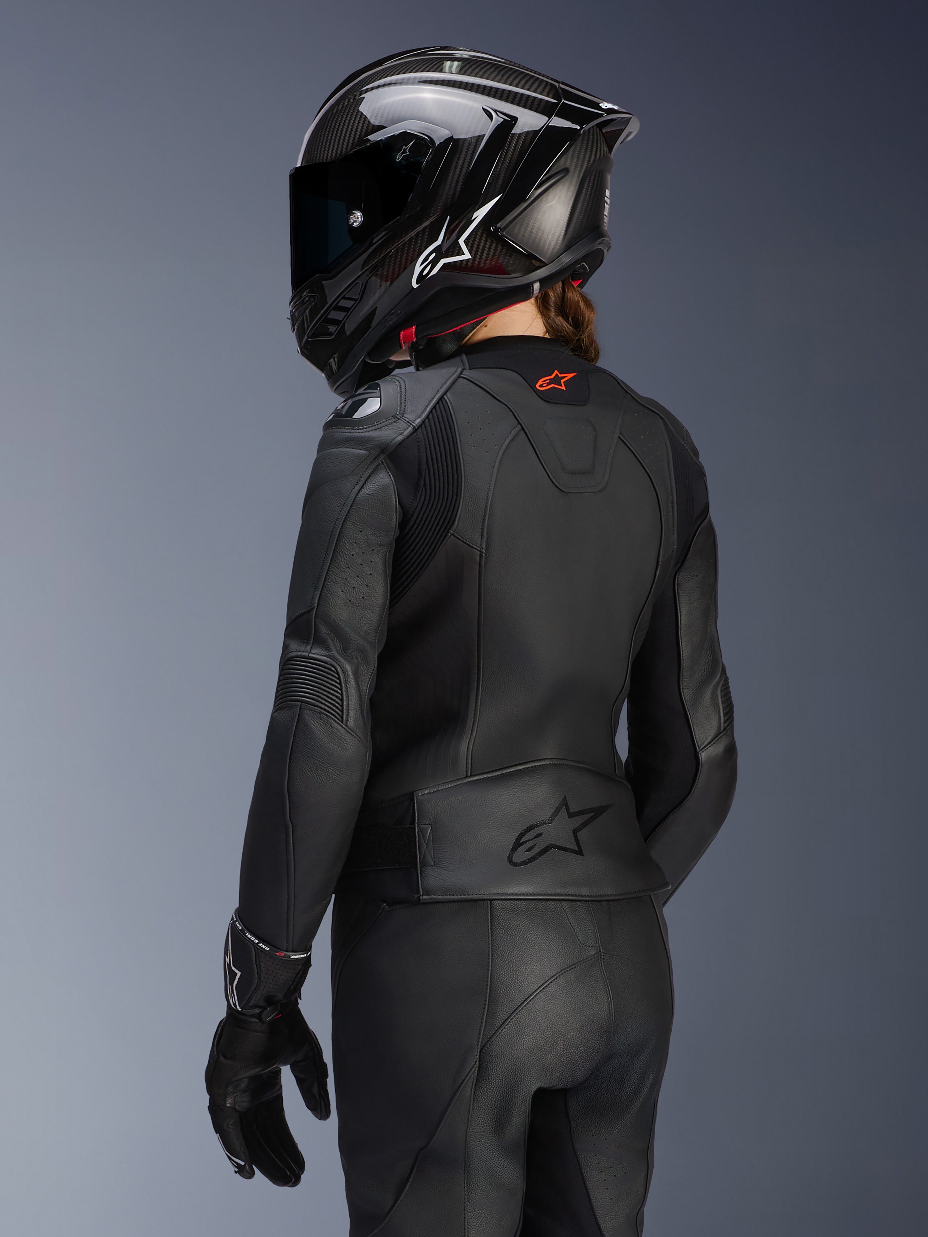 Rückansicht einer Fahrerin in der Alpinestars Stella GP Plus V4 Damen-Lederjacke in Schwarz, mit passender Lederhose, Handschuhen und einem schwarzen Integralhelm im Carbon-Stil; hervorgehoben werden die aerodynamische Passform der Jacke und das geprägte Logo am unteren Rücken.