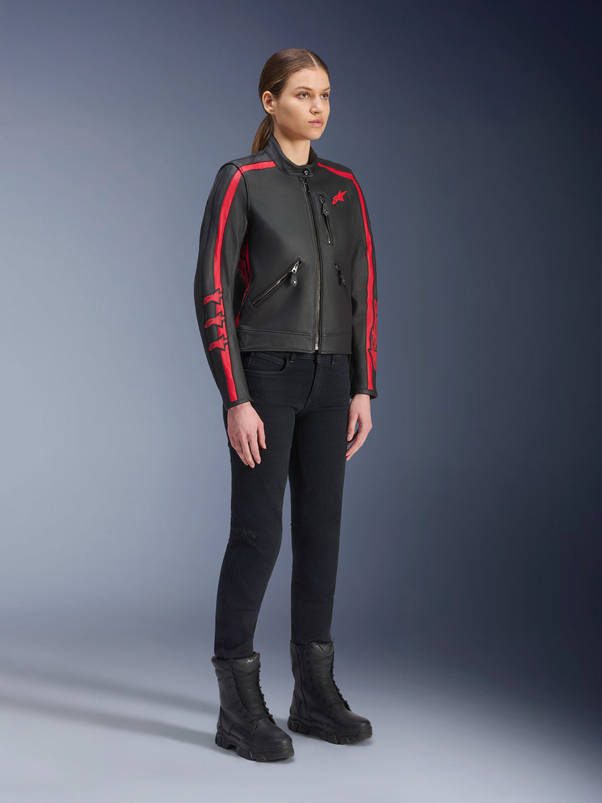 Weibliches Model in der Alpinestars Stella Dyno Lederjacke in Black Haute Red, kombiniert mit schwarzer Hose und Stiefeln, Motorrad-Lederjacke im Heritage-Stil mit roten Streifen an den Armen und rotem Logo, urbane Motorradausrüstung für Damen