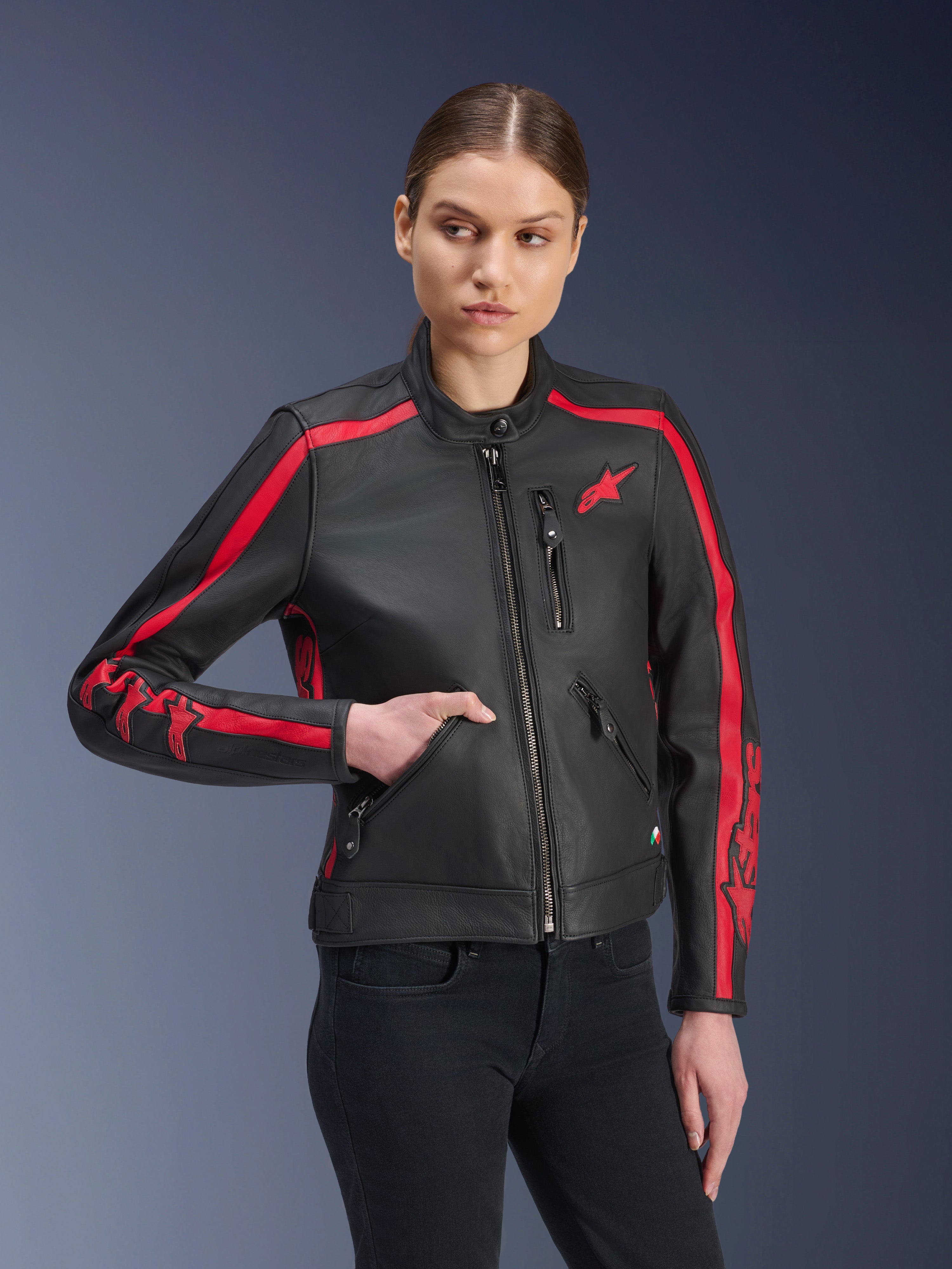 Alpinestars Stella Dyno Lederjacke, Damen-Motorradjacke, Schwarz Haute Red, von einem Modell mit schwarzen Jeans getragen, mit roten Rennstreifen an Schultern und Ärmeln sowie markantem Branding, hochwertige Lederkonstruktion, Heritage-inspiriertes Design für urbanes Fahren