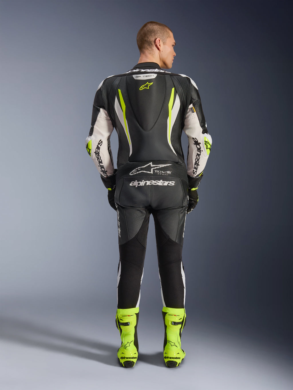 Alpinestars GP Tech V5 Einteiliger Lederkombi, Rückansicht, Schwarz, Weiß und Gelb Fluo, mit aerodynamischem Rückenhöcker mit gelbem Stern-Logo, weißes Alpinestars-Branding an Ärmeln und Rücken, passende neongelbe Rennstiefel, professionelle Motorrad-Rennausrüstung