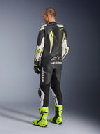 GP Tech V5 Einteiliger Lederanzug, Schwarz, Weiß und Gelb Fluo, Rückansicht eines Fahrers in vollständigem Rennanzug mit passenden neongelben Stiefeln, mit aerodynamischem Rückenhöcker und markanten Alpinestars-Logos, professionelle Motorradausrüstung