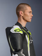 Männliches Model im Alpinestars GP Tech V5 Einteiler-Lederanzug in Schwarz-Weiß-Gelb Fluo, Seitenansicht mit Fokus auf den aerodynamischen Rückenhöcker und die weißen Schulter-Slider, rennsportinspirierte Motorrad-Schutzausrüstung mit perforierten Lederdetails