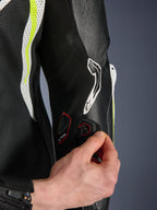 Alpinestars GP Tech V5 Einteiliger Lederkombi, Detailansicht des Tech-Air Anschlussports am Ärmel, Hand demonstriert elektronische Integration, schwarzes Leder mit gelben Fluo- und weißen perforierten Einsätzen, professionelle Rennausrüstung