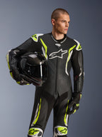 Alpinestars GP Tech V5 Einteiliger Lederkombi, Schwarz, Weiß und Gelb Fluo, männliches Model mit schwarzem Motorradhelm, mit perforierten Panels, weißen und neongelben Akzenten sowie markantem Alpinestars-Branding, professionelle Motorrad-Rennausrüstung