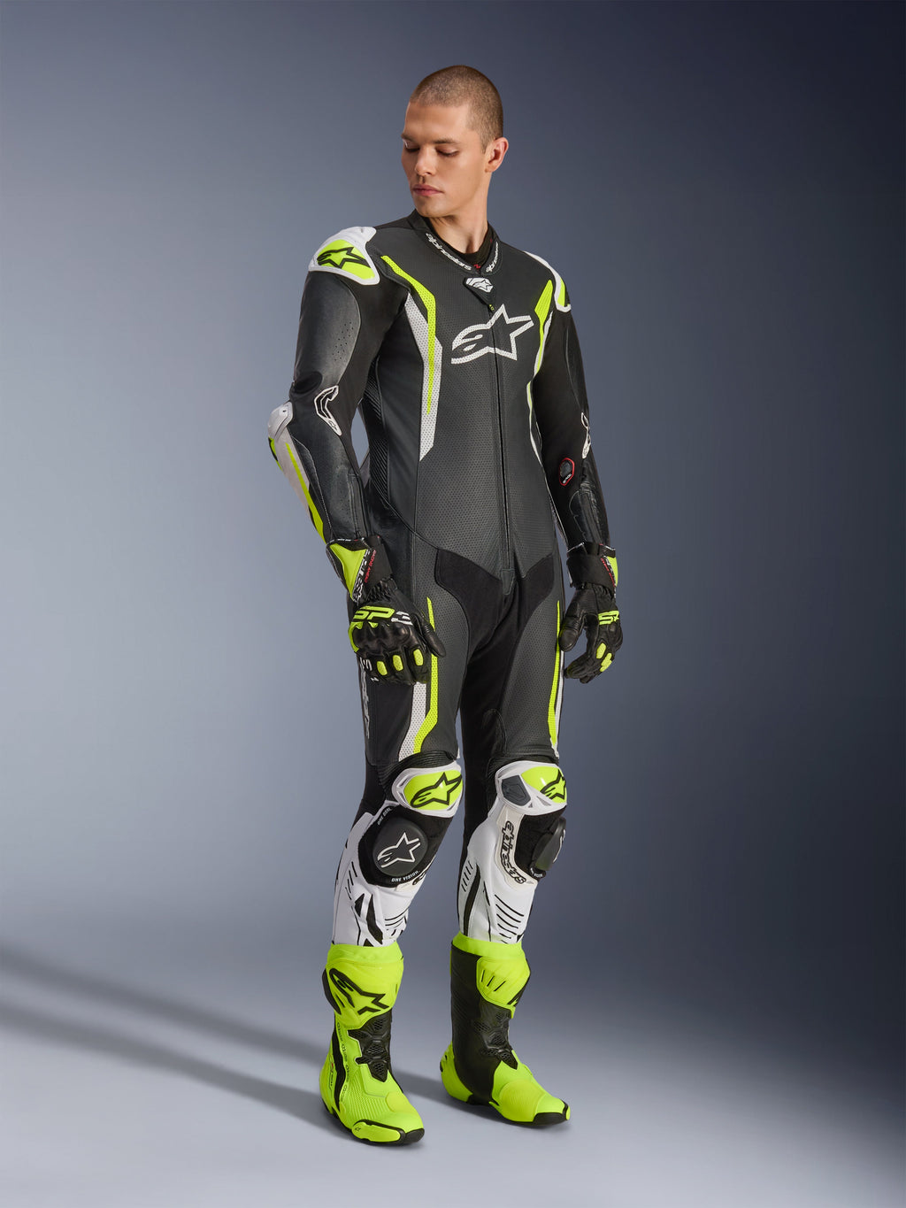 Alpinestars GP Tech V5 Einteiliger Lederkombi, Schwarz, Weiß und Gelb Fluo, getragen von einem Model mit passenden Rennhandschuhen und Stiefeln, professionelle Motorrad-Rennausrüstung mit aerodynamischer Performance und Schutzausrüstung.