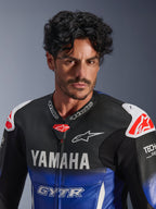 Close-up portrait of a man wearing the Alpinestars Yamaha GP-R7 One Piece Leather Suit, Metallic Blue and Black with white YAMAHA and GYTR branding, perforated leather, white shoulder sliders with red star logos, Tech-Air Ready, professional motorcycle racing gear.