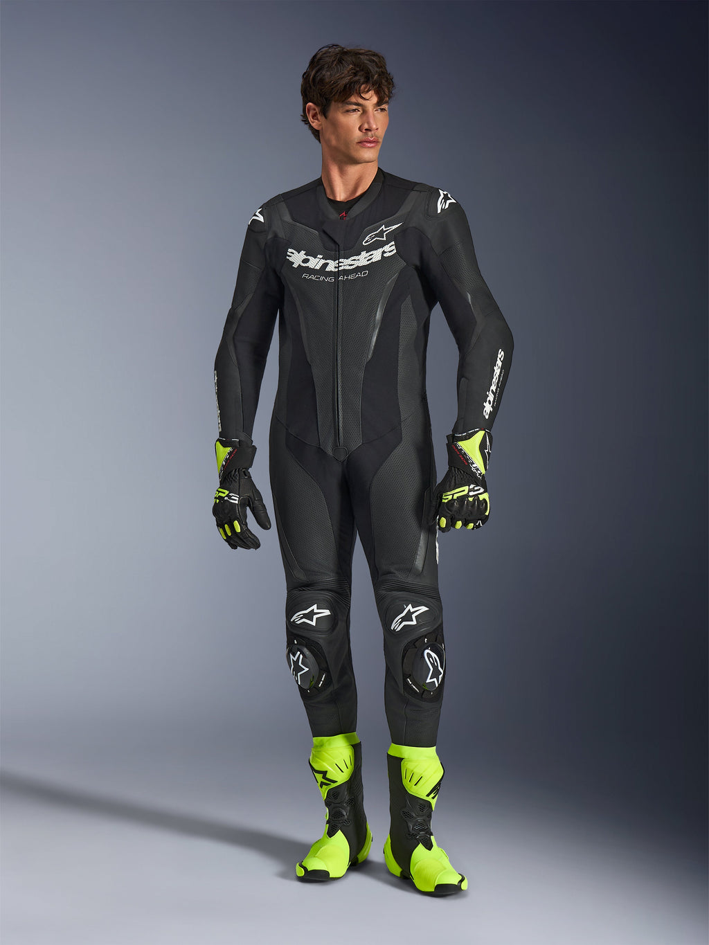 Alpinestars GP Force V2 Lederkombi, einteiliger schwarzer Motorradrennanzug, getragen mit neongelben Stiefeln und Handschuhen, mit weißen Logos und perforiertem Leder, schlankes Design für Trackdays