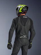 Alpinestars GP Force V2 Lederkombi 1-teilig, Schwarz, Rückansicht mit aerodynamischem Speed-Hump und perforierter Lederkonstruktion, getragen mit einem schwarz-neongelben Rennhelm und schwarzen Handschuhen, mit Akkordeon-Stretcheinsätzen und weißem Tech-Air Ready-Branding.