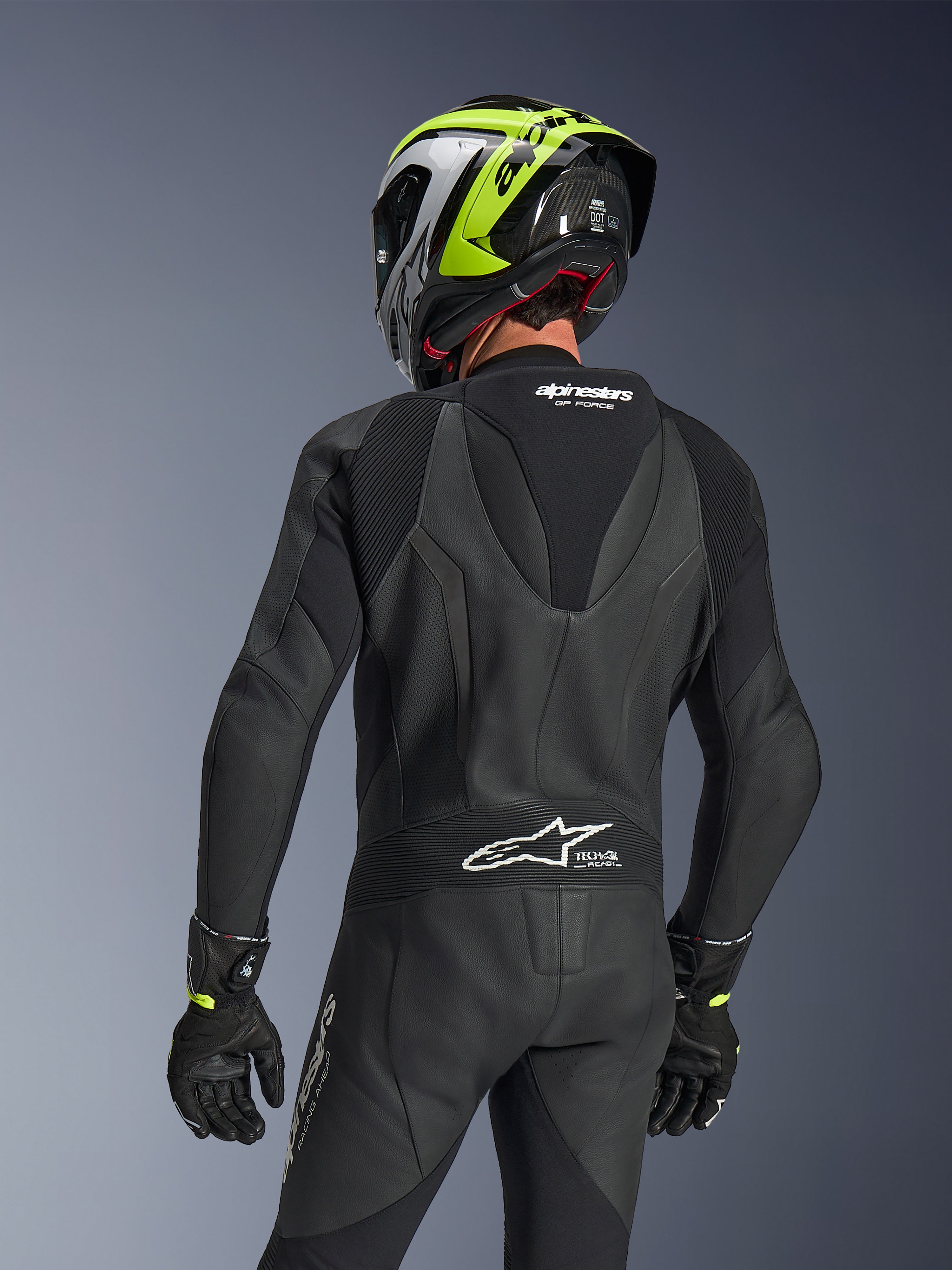 Alpinestars GP Force V2 Lederkombi 1-teilig, Schwarz, Rückansicht mit aerodynamischem Speed-Hump und perforierter Lederkonstruktion, getragen mit einem schwarz-neongelben Rennhelm und schwarzen Handschuhen, mit Akkordeon-Stretcheinsätzen und weißem Tech-Air Ready-Branding.