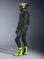 Motorradfahrer im Alpinestars GP Force V2 Lederkombi 1-teilig in Schwarz, Rückansicht mit weißem Branding am Oberschenkel und unteren Rücken, aerodynamischer Rückenhöcker, kombiniert mit einem neongelb-schwarzen Helm, passenden Rennhandschuhen und Stiefeln, professionelle Rennstrecken- und Sportfahrausrüstung.