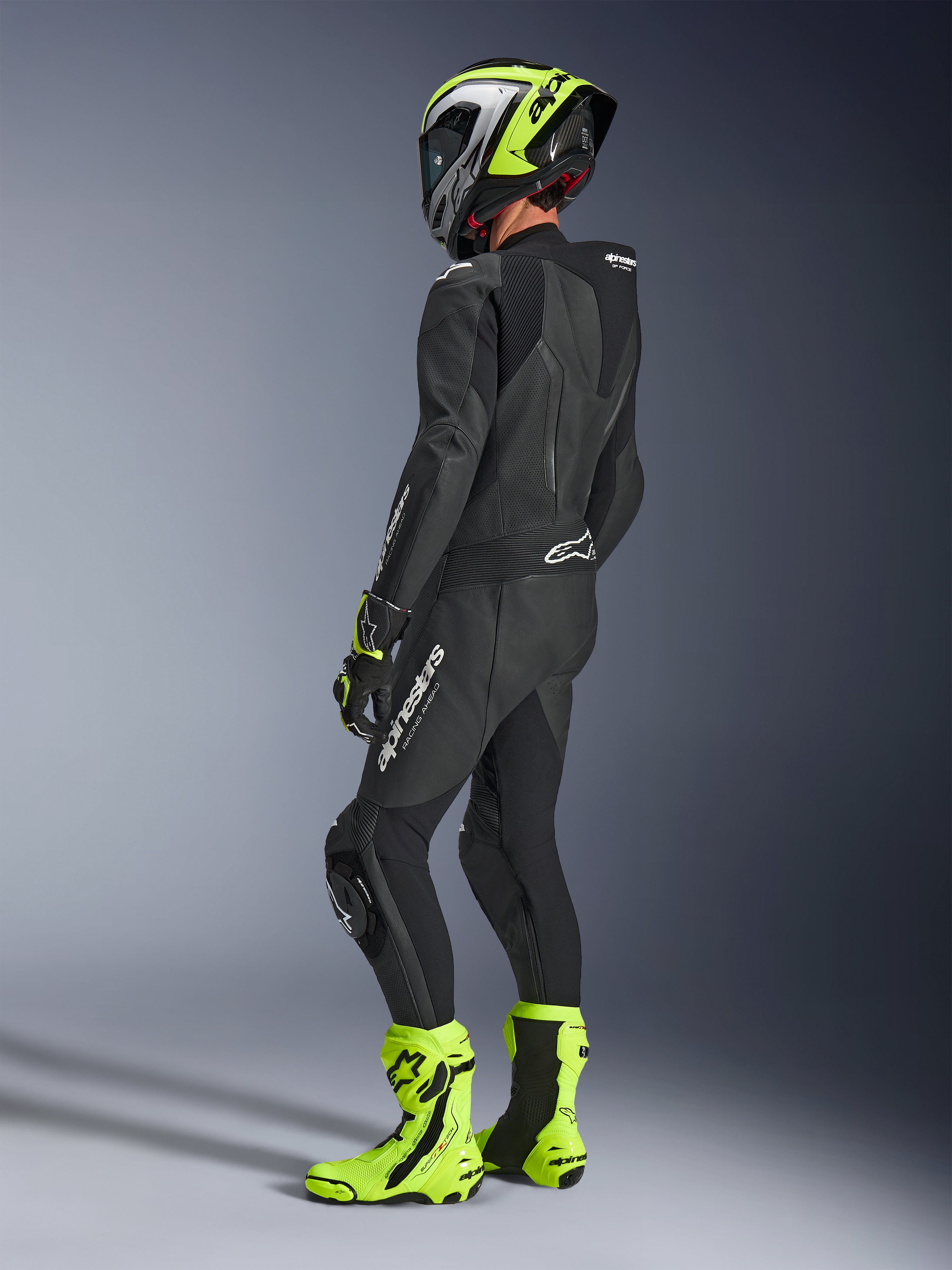 Motorradfahrer im Alpinestars GP Force V2 Lederkombi 1-teilig in Schwarz, Rückansicht mit weißem Branding am Oberschenkel und unteren Rücken, aerodynamischer Rückenhöcker, kombiniert mit einem neongelb-schwarzen Helm, passenden Rennhandschuhen und Stiefeln, professionelle Rennstrecken- und Sportfahrausrüstung.
