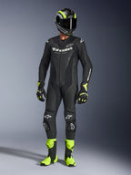 Motorradfahrer im Alpinestars GP Force V2 Lederanzug in Schwarz, einteiliger Lederanzug mit weißem Branding, kombiniert mit einem schwarz-fluorgelben Rennhelm, Handschuhen und Stiefeln, professionelle Rennausrüstung auf grauem Studiohintergrund