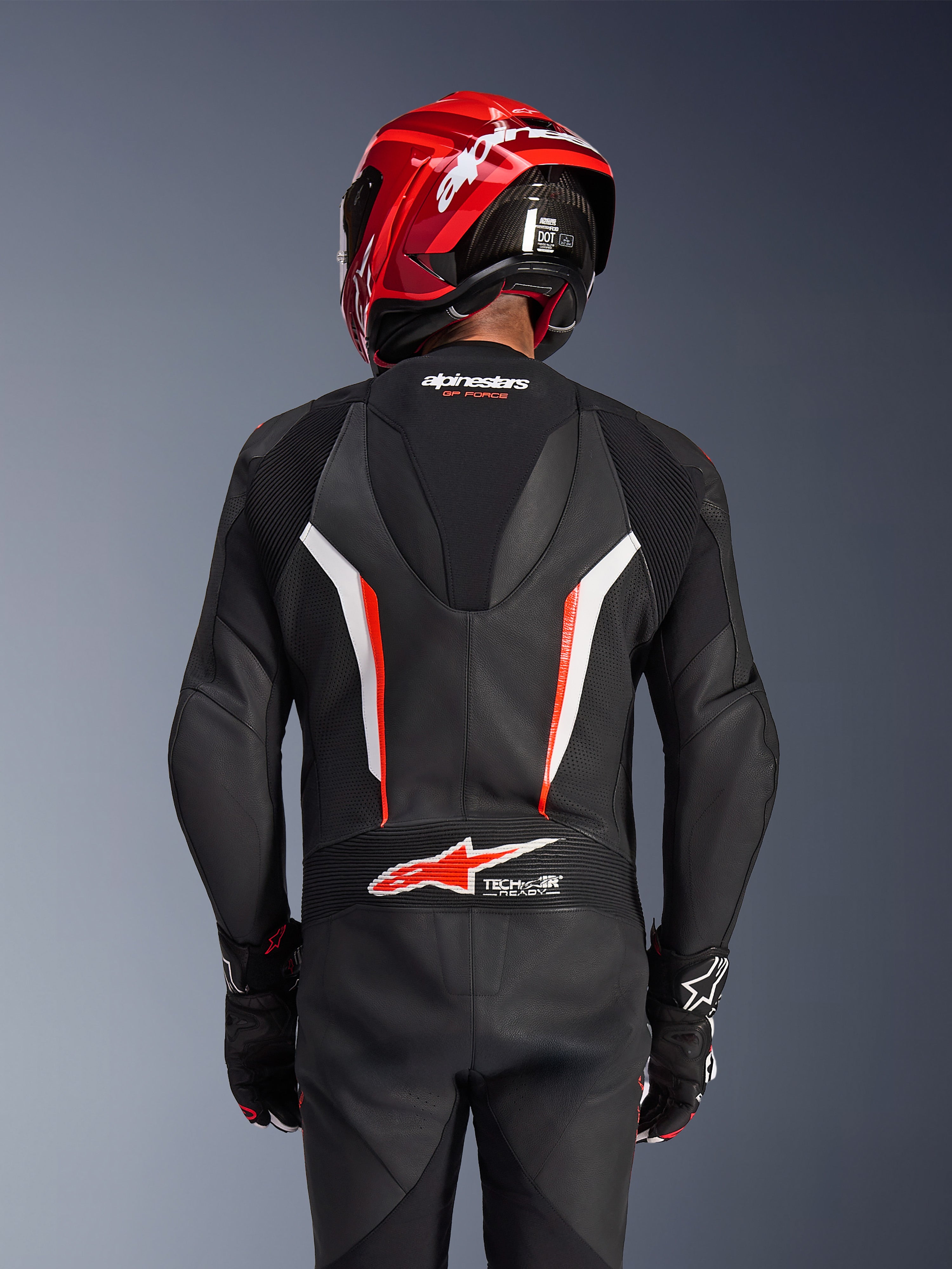 Alpinestars GP Force V2 1-Teiler Lederkombi, Schwarz, Weiß und Rot Fluo, Rückansicht eines Fahrers mit rotem Rennhelm und Handschuhen, mit perforierten Lederbahnen, aerodynamischem Rückenhöcker mit Alpinestars-Branding und Tech-Air Ready-Logo am unteren Rücken, professionelle Motorrad-Rennausrüstung.