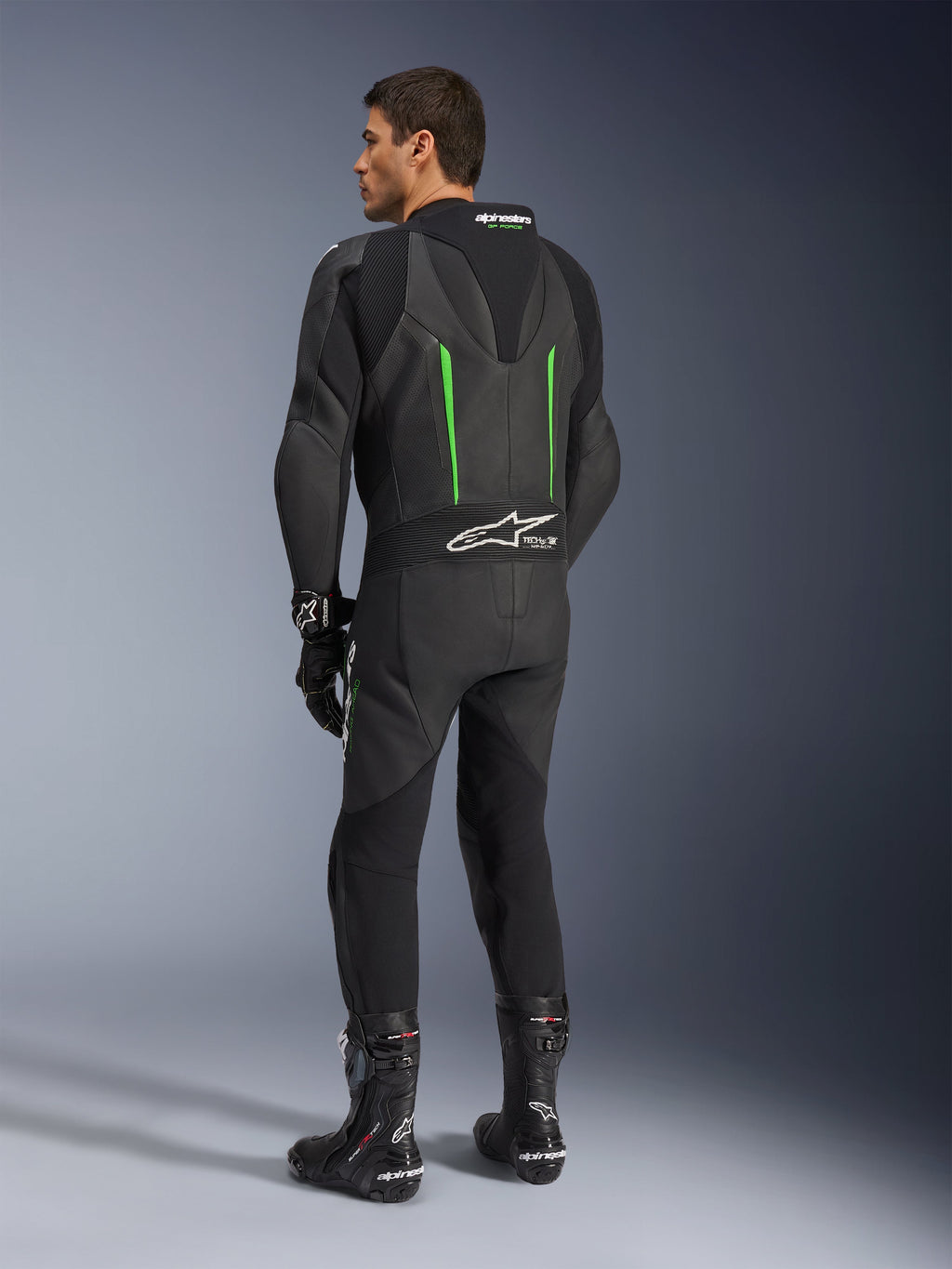 Alpinestars GP Force V2 Lederkombi, einteiliger Motorradanzug, Schwarz mit grünen Akzenten, Rückansicht mit aerodynamischem Rückenhöcker und weißem Logo auf dem Akkordeon-Stretcheinsatz, kombiniert mit schwarzen Rennstiefeln und Handschuhen.