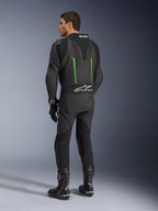 Alpinestars GP Force V2 Lederkombi, einteiliger Motorradanzug, Schwarz mit grünen Akzenten, Rückansicht mit aerodynamischem Rückenhöcker und weißem Logo auf dem Akkordeon-Stretcheinsatz, kombiniert mit schwarzen Rennstiefeln und Handschuhen.