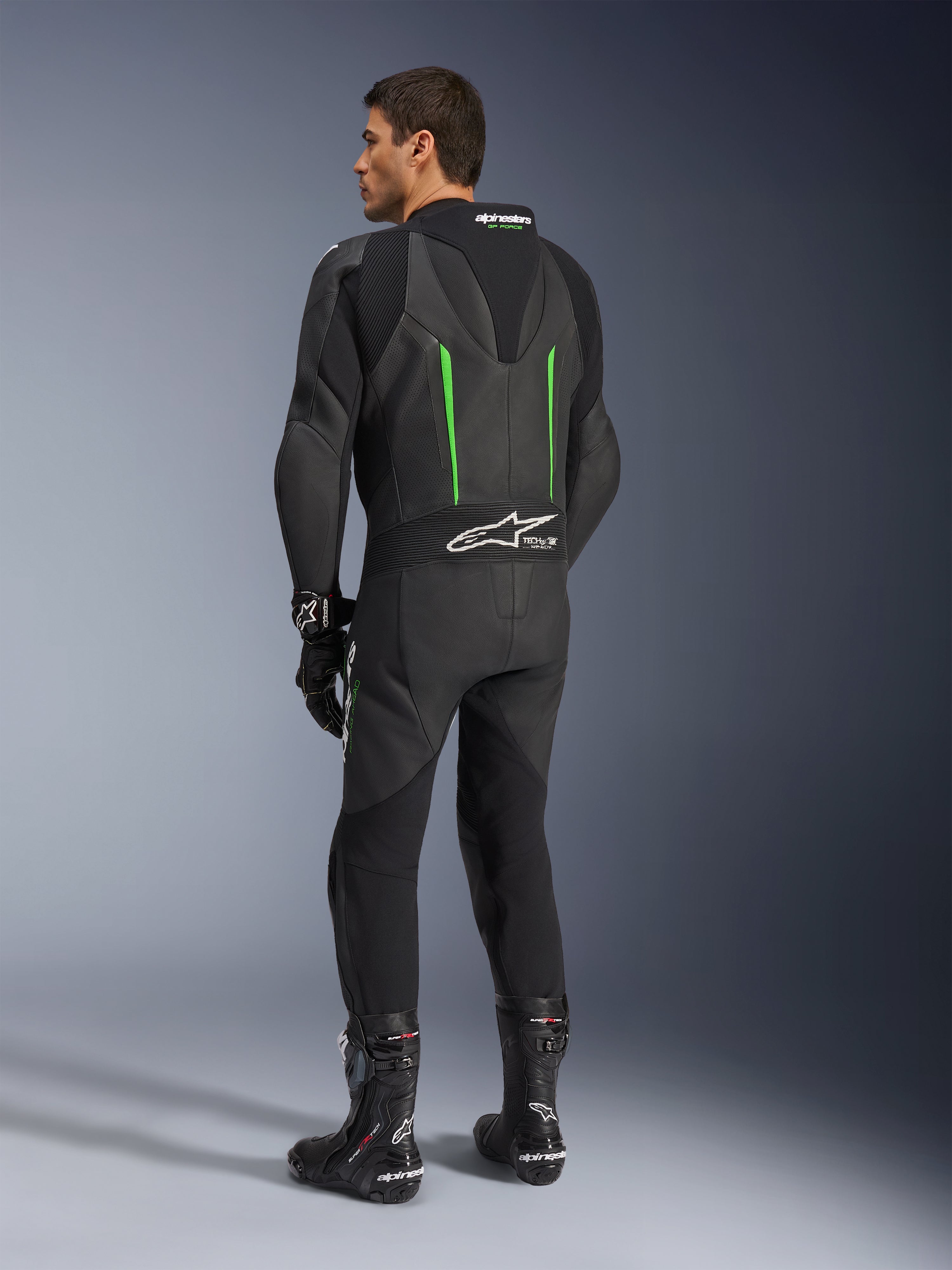 Alpinestars GP Force V2 Lederkombi, einteiliger Motorradanzug, Schwarz mit grünen Akzenten, Rückansicht mit aerodynamischem Rückenhöcker und weißem Logo auf dem Akkordeon-Stretcheinsatz, kombiniert mit schwarzen Rennstiefeln und Handschuhen.