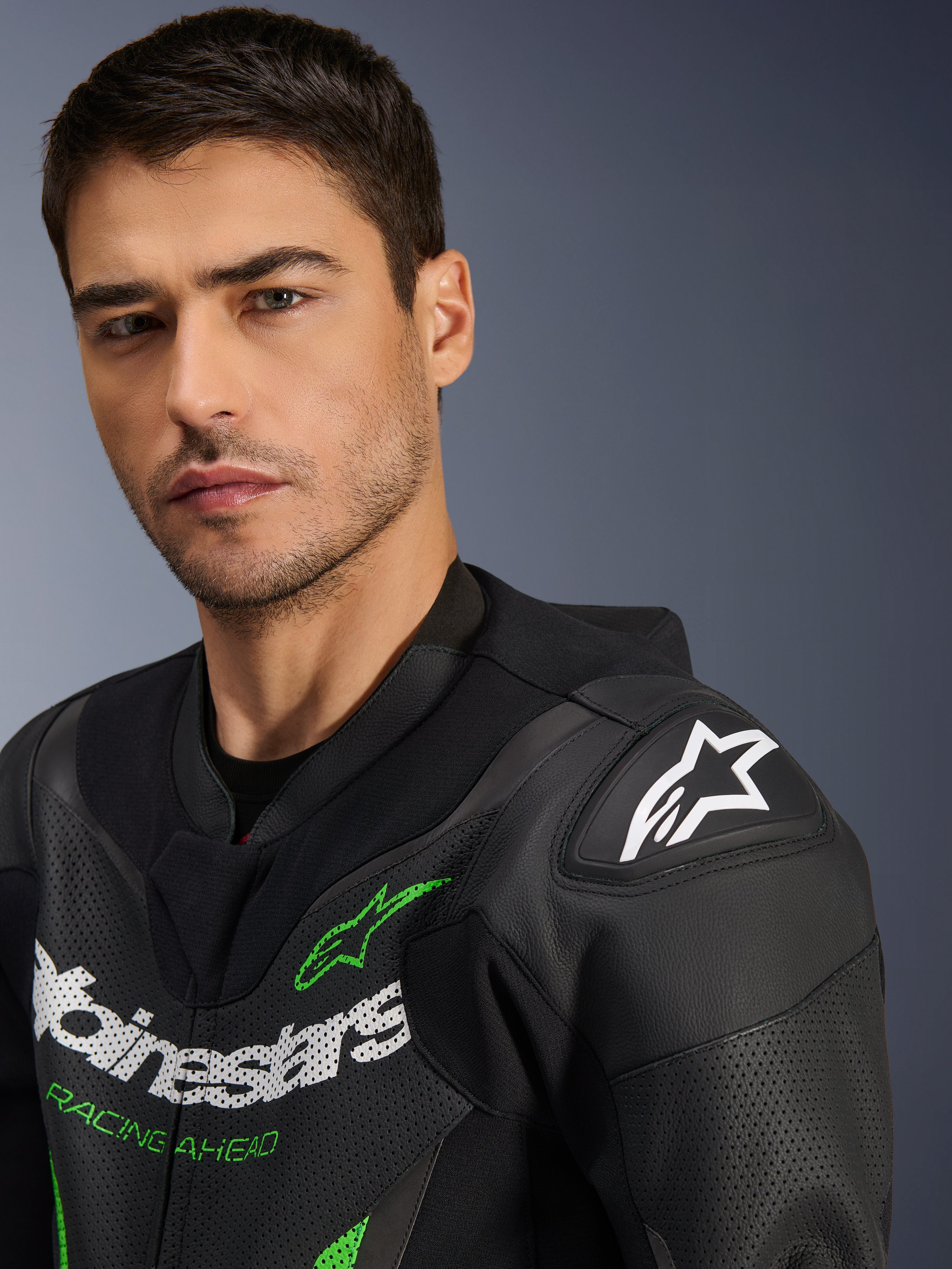Alpinestars GP Force V2 Lederkombi – 1-teilig, Schwarz und Grün, Nahaufnahme eines männlichen Models im Motorrad-Rennanzug, mit weißem und grünem Logo-Branding, perforierten Lederpaneelen und DFS Lite Schulterprotektoren mit weißem Stern-Logo.