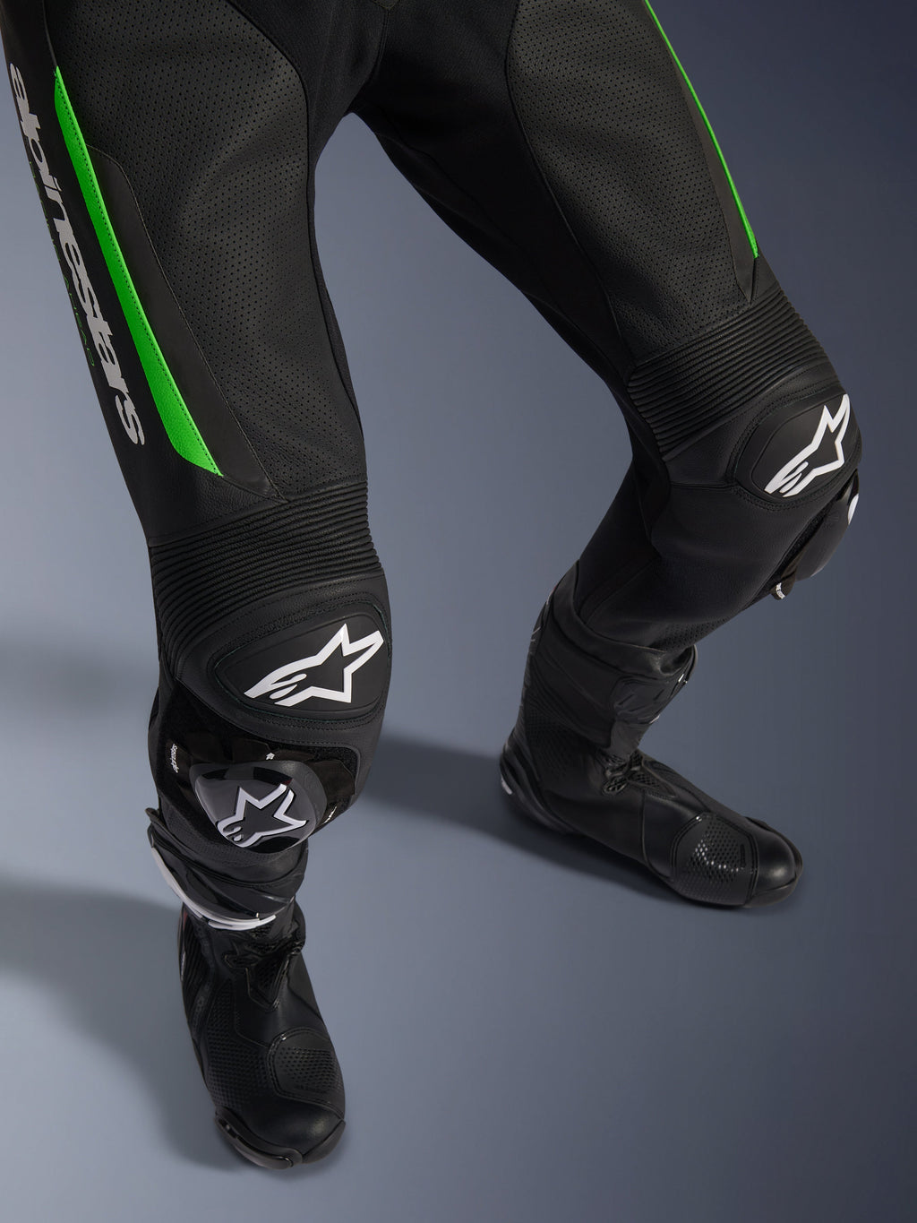 Alpinestars GP Force V2 Lederkombi - 1-teilig, Detailaufnahme der Beine in Schwarz und Grün, mit perforierter Lederkonstruktion, Akkordeon-Stretch-Einsätzen an den Knien, weißem Alpinestars-Branding und integrierten Knieschleifern, getragen mit schwarzen Rennstiefeln