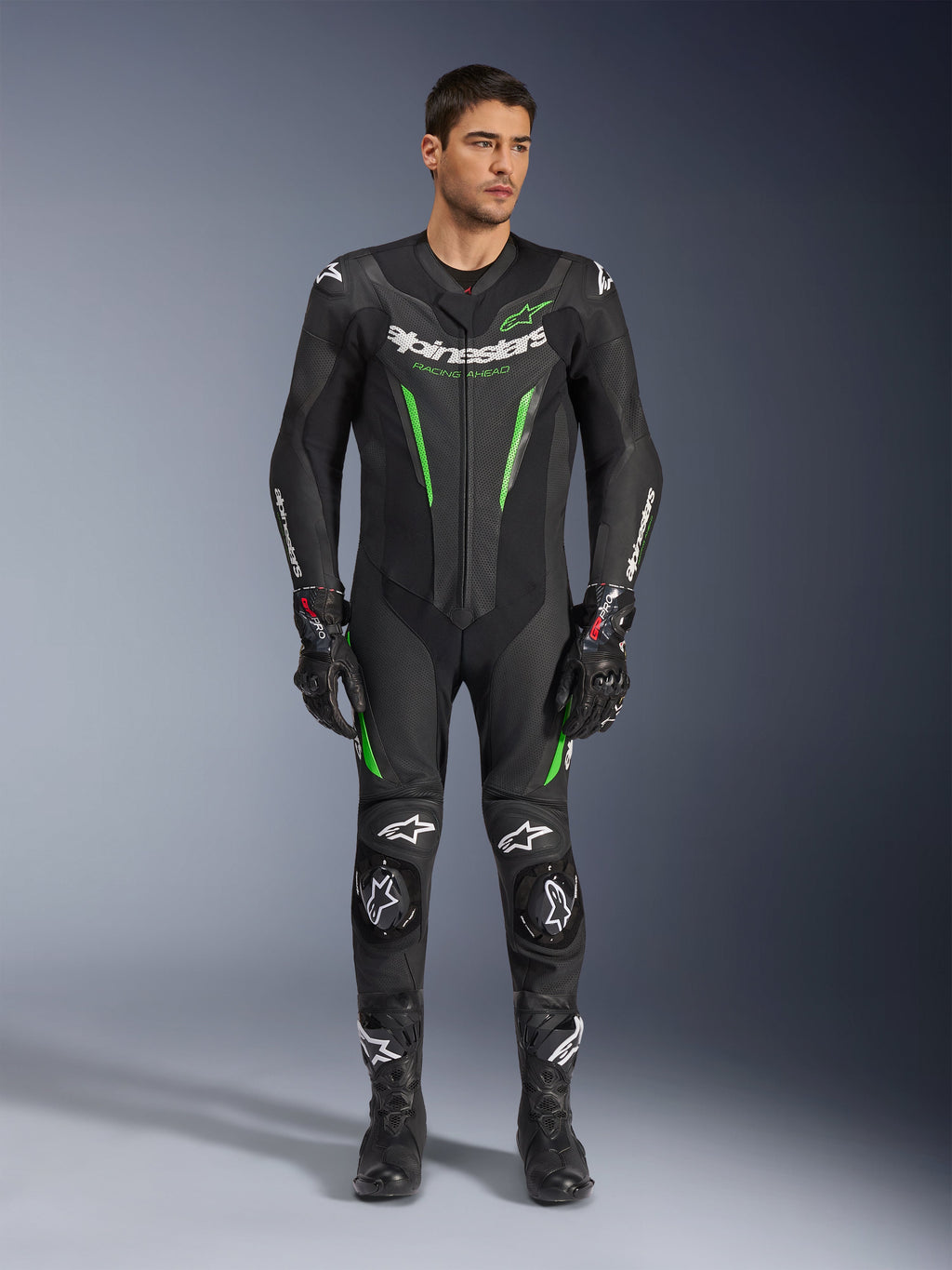 Alpinestars GP Force V2 Lederkombi – 1-teilig, einteiliger Motorrad-Rennanzug, Schwarz und Grün, Frontansicht eines Models im Anzug mit weißem Branding auf der Brust und neongrünen Akzenten, mit perforiertem Leder und externen Schulterschleifern, kombiniert mit schwarzen Rennhandschuhen und Stiefeln