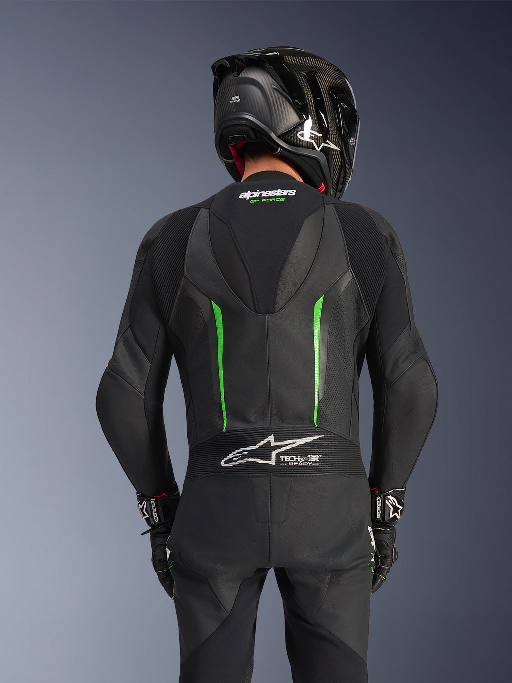 Rückansicht eines Fahrers im Alpinestars GP Force V2 Lederkombi in Schwarz-Grün, mit aerodynamischem Höcker, Tech-Air Ready-Branding auf dem Akkordeon-Einsatz am unteren Rücken und perforierter Lederkonstruktion, kombiniert mit einem schwarzen Carbon-Rennhelm.