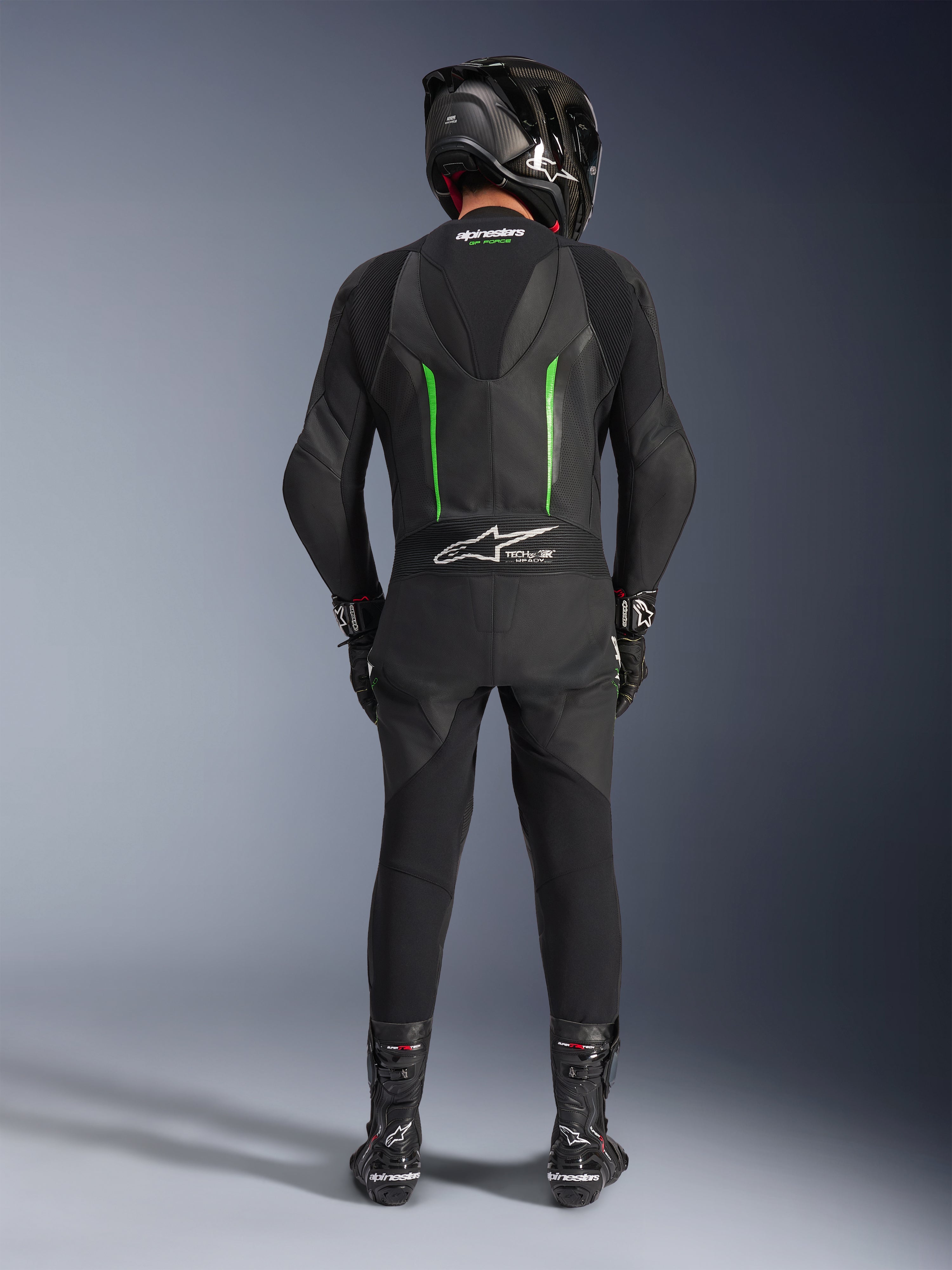 Fahrer in der Rückansicht im Alpinestars GP Force V2 Lederanzug – Einteiler in Schwarz und Grün, kombiniert mit einem schwarzen Carbon-Helm, schwarzen Rennsport-Handschuhen und schwarzen Stiefeln, mit aerodynamischem Geschwindigkeitsbuckel und Tech-Air Ready-Branding.