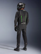 Fahrer von hinten im Alpinestars GP Force V2 Lederkombi, Einteiler, Schwarz mit grünen Akzenten, aerodynamischer Rückenhöcker, kombiniert mit einem schwarzen Carbon-Helm, schwarzen Rennstiefeln und Handschuhen, Hochleistungs-Motorrad-Rennausrüstung