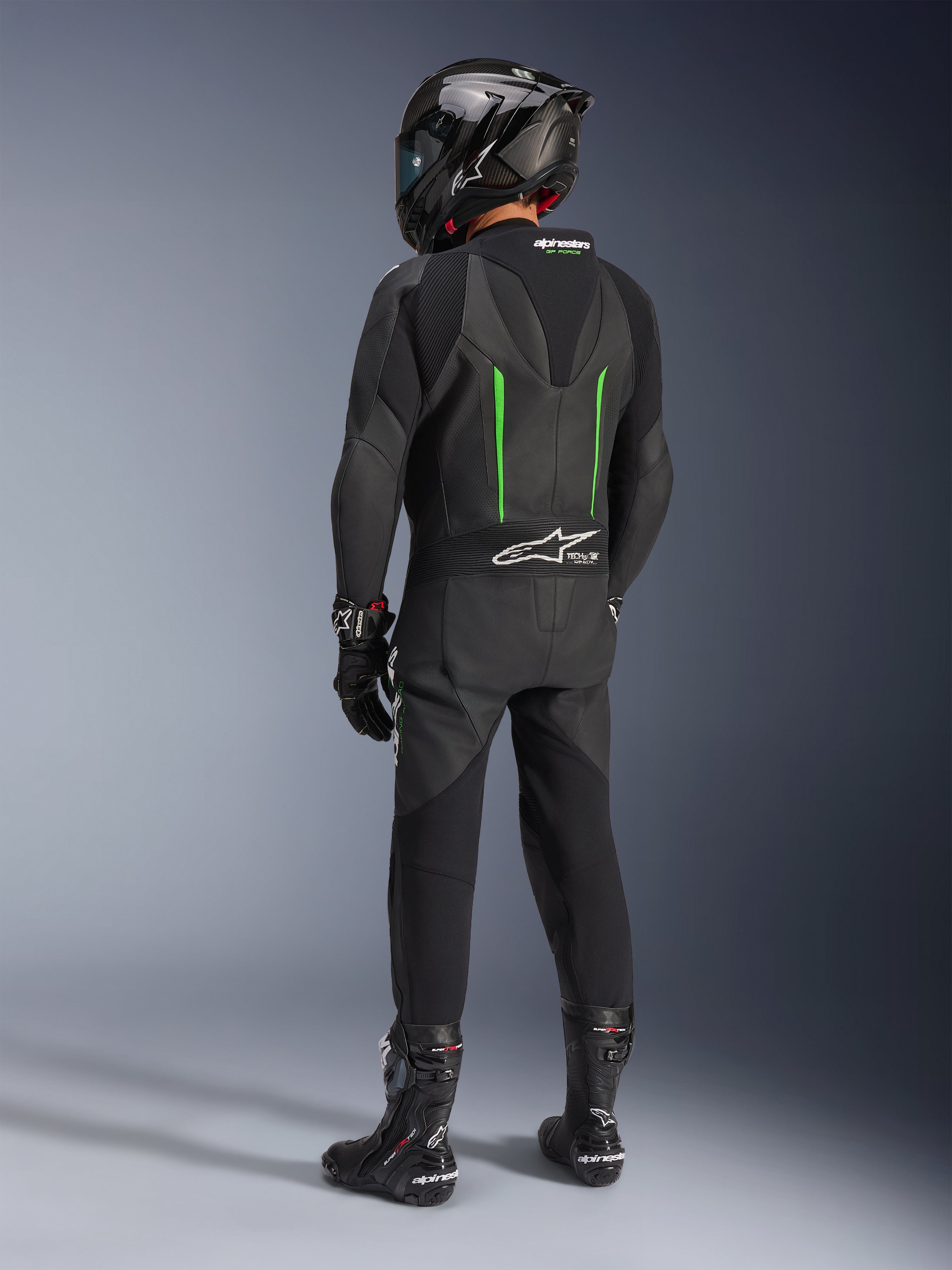 Fahrer von hinten im Alpinestars GP Force V2 Lederkombi, Einteiler, Schwarz mit grünen Akzenten, aerodynamischer Rückenhöcker, kombiniert mit einem schwarzen Carbon-Helm, schwarzen Rennstiefeln und Handschuhen, Hochleistungs-Motorrad-Rennausrüstung