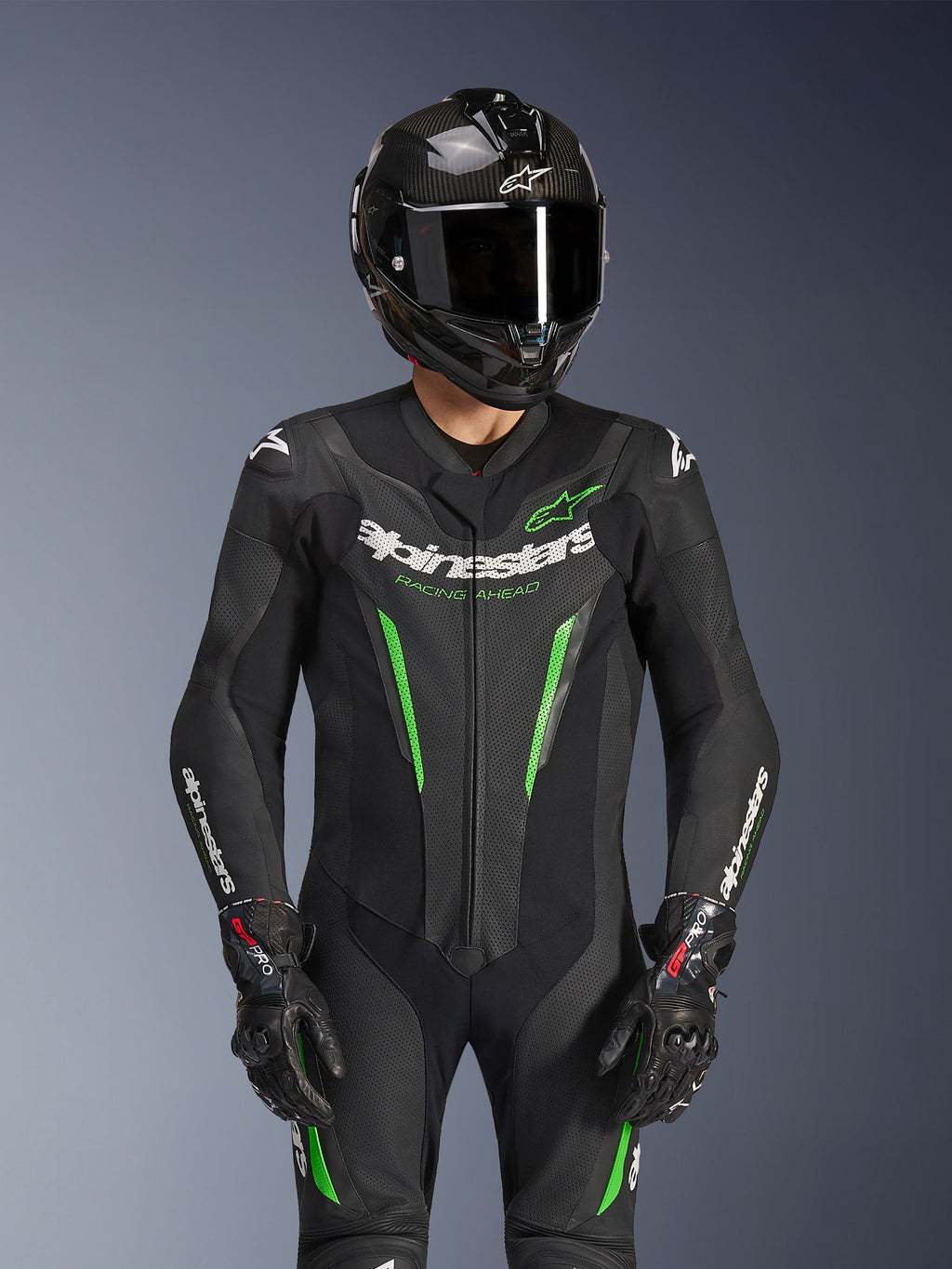 Alpinestars GP Force V2 Lederkombi – 1-teilig, Schwarz und Grün, Motorrad-Rennanzug getragen mit einem Carbonfaser-Helm und GP Pro Handschuhen, mit limettengrünen Akzenten und Alpinestars „Racing Ahead“-Branding, entwickelt für Rennstrecken-Performance.