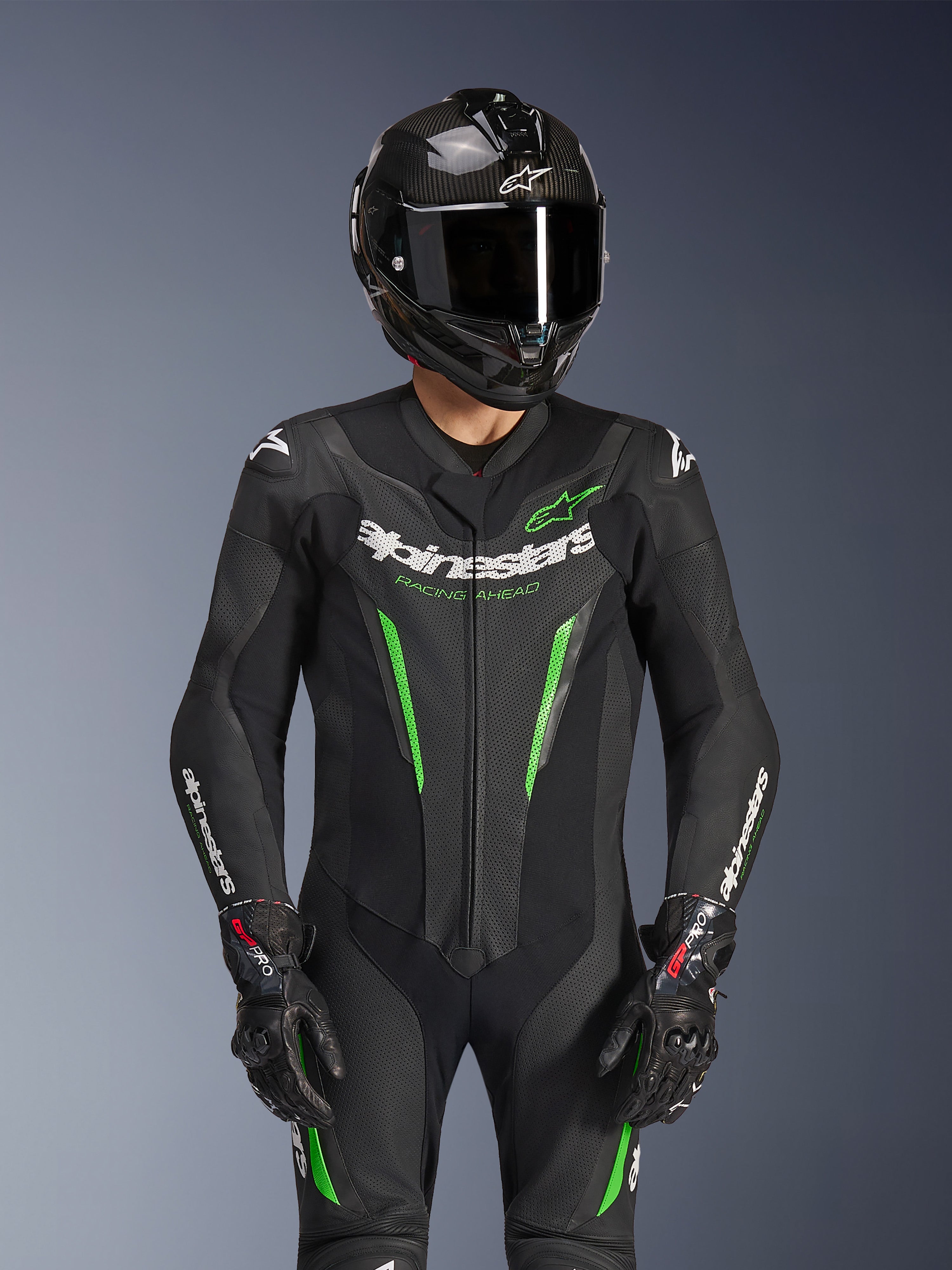 Alpinestars GP Force V2 Lederkombi – 1-teilig, Schwarz und Grün, Motorrad-Rennanzug getragen mit einem Carbonfaser-Helm und GP Pro Handschuhen, mit limettengrünen Akzenten und Alpinestars „Racing Ahead“-Branding, entwickelt für Rennstrecken-Performance.