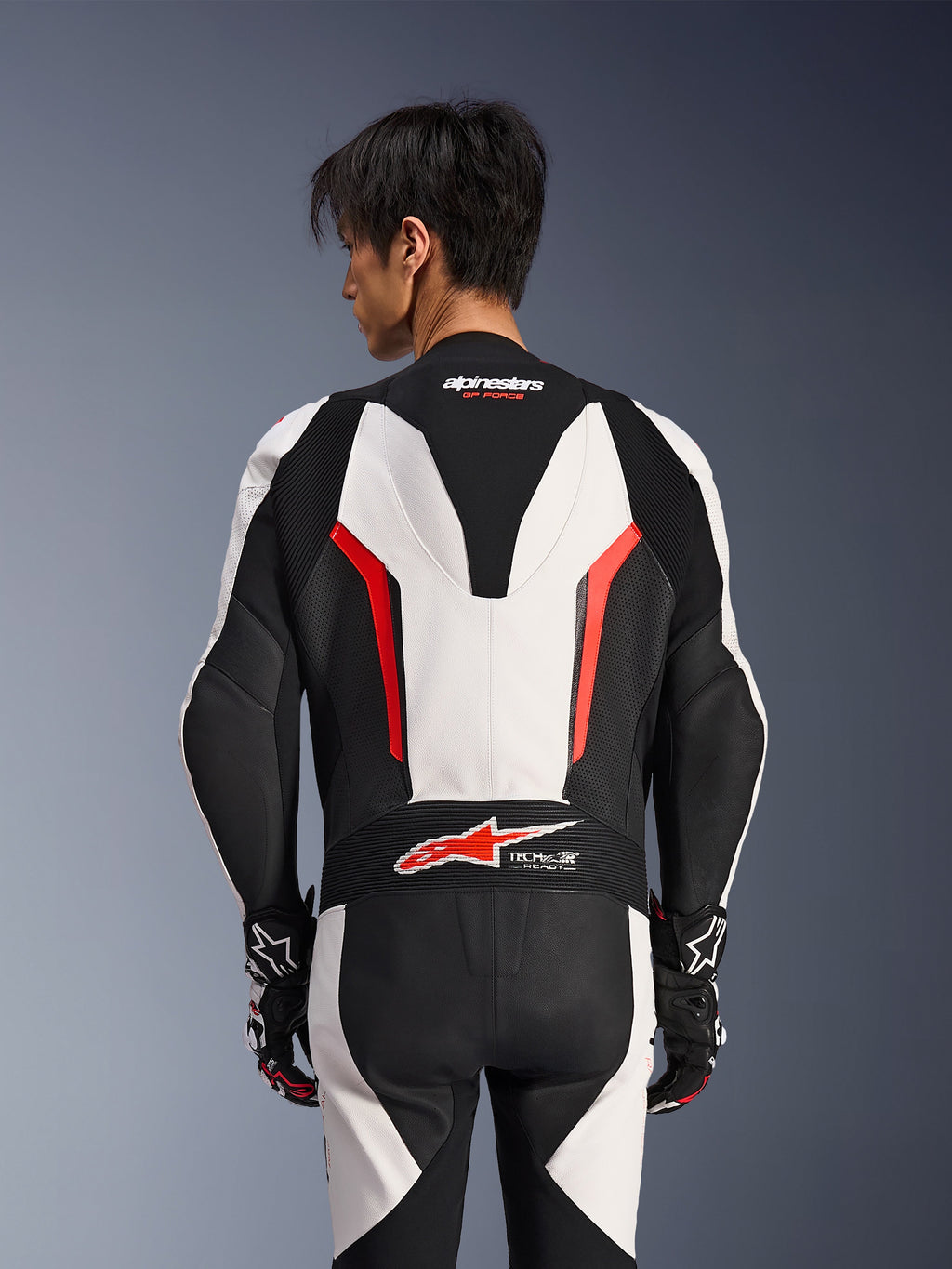 Alpinestars GP Force V2 Lederkombi 1-teilig, Weiß, Schwarz und Rot Fluo, Rückansicht an einem männlichen Model, mit aerodynamischem Höcker, perforierten Lederbahnen und Tech-Air Ready-Logo am unteren Rücken, professionelle Rennausrüstung mit schwarz-weißen Handschuhen