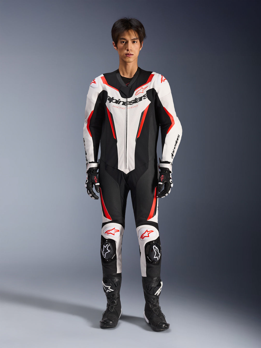 Alpinestars GP Force V2 Lederkombi - 1-teilig, Weiß Schwarz Rot Fluo, einteiliger Motorrad-Lederkombi, Vorderansicht am Modell, mit perforierten Einsätzen und roten Akzenten, markantes Alpinestars-Branding auf der Brust, kombiniert mit professionellen Rennstiefeln und Handschuhen