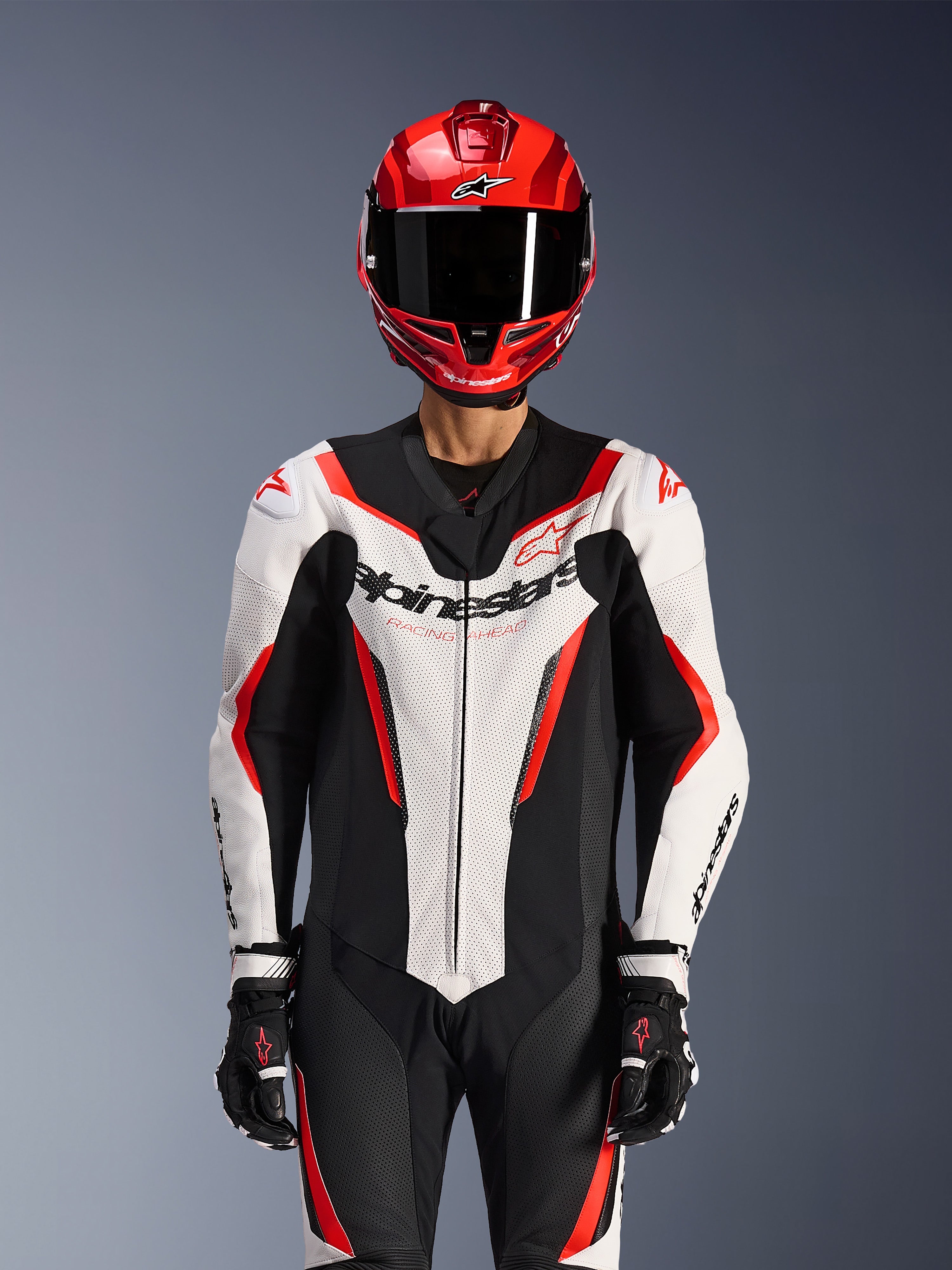 Alpinestars GP Force V2 Lederkombi - 1-teilig, Weiß Schwarz Rot Fluo, getragen mit rotem Integralhelm und schwarzen Rennhandschuhen, mit perforierten Lederbahnen und markantem Alpinestars-Branding auf der Brust, professionelle Performance-Ausrüstung für Motorradrennen