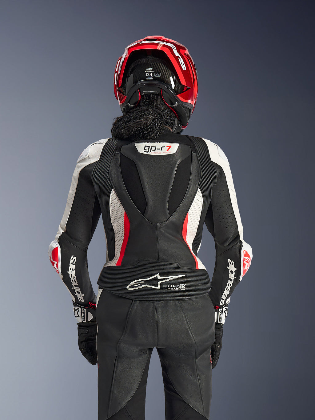 Alpinestars Stella GP-R7 2-Teiler-Lederanzug für Damen, Schwarz, Weiß und Fluo-Rot, Rückansicht einer Fahrerin mit rotem Helm und schwarzen Handschuhen, aerodynamischer Höcker mit GP-R7-Branding und Tech-Air Ready-Logo am unteren Rücken.