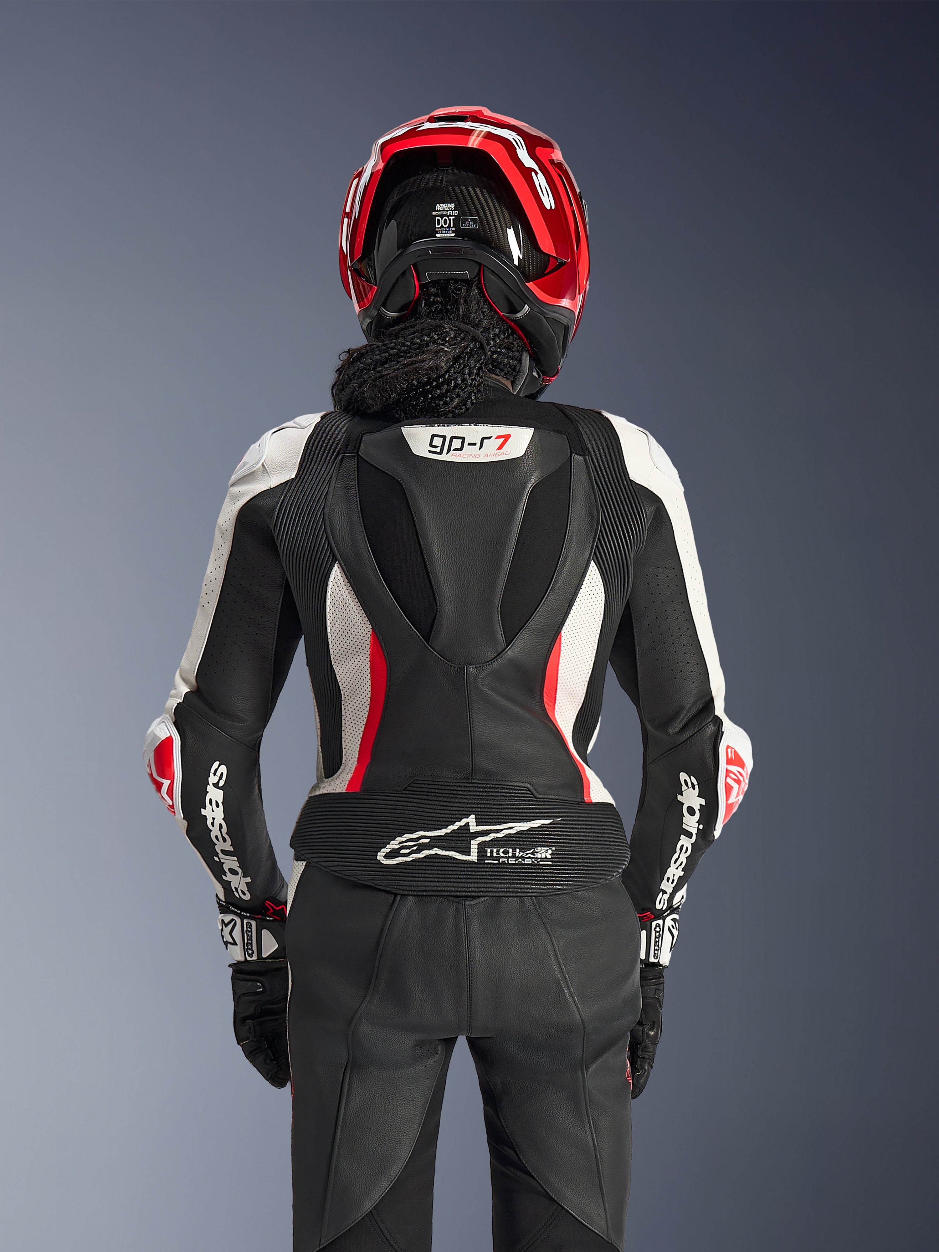 Alpinestars Stella GP-R7 2-Teiler-Lederanzug für Damen, Schwarz, Weiß und Fluo-Rot, Rückansicht einer Fahrerin mit rotem Helm und schwarzen Handschuhen, aerodynamischer Höcker mit GP-R7-Branding und Tech-Air Ready-Logo am unteren Rücken.