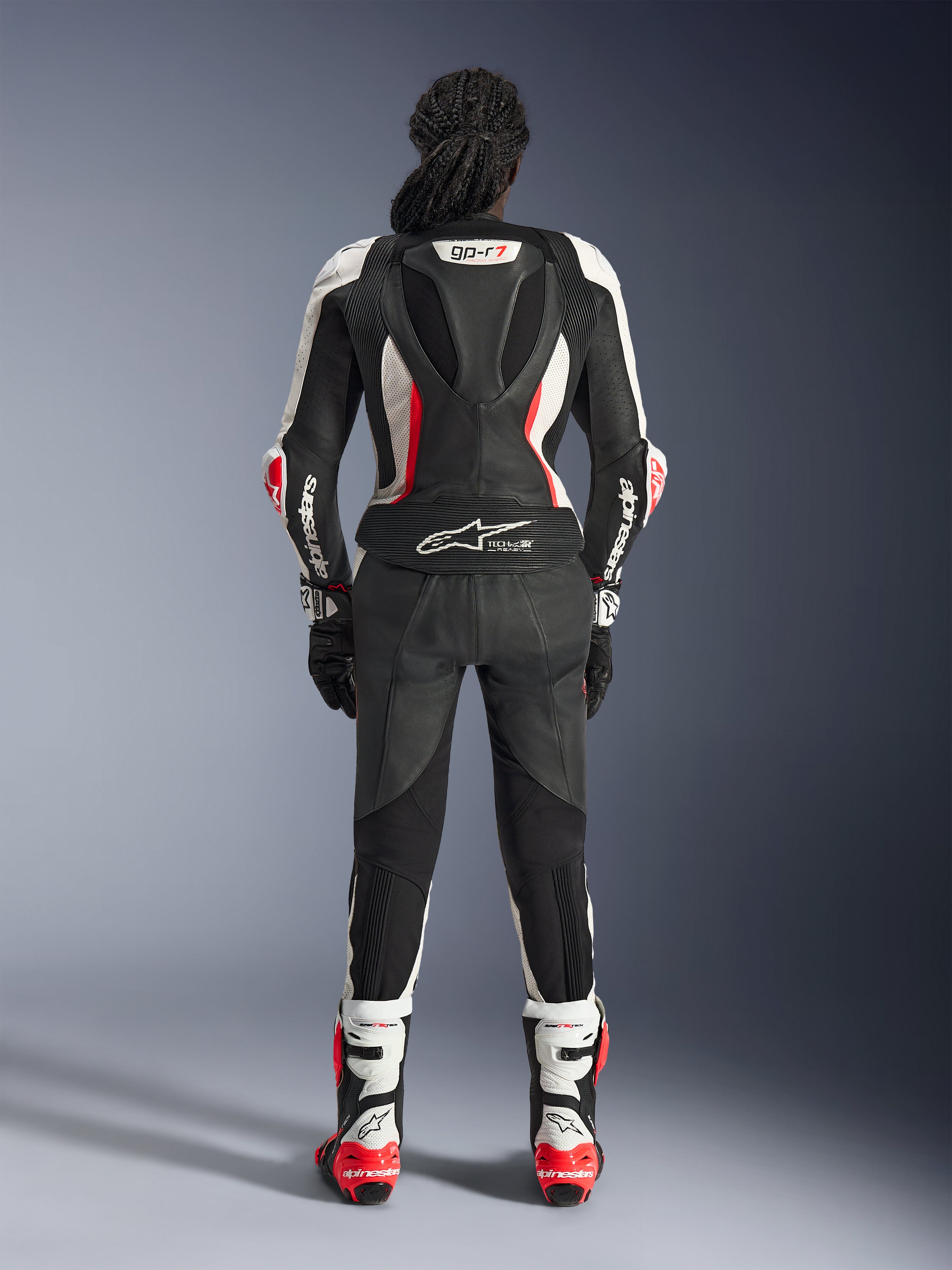 Rückansicht des Alpinestars Stella GP-R7 2-teiligen Lederanzugs, Schwarz, Weiß und Fluo-Rot, mit einem aerodynamischen Höcker samt GP-R7-Branding und Tech-Air Ready-Logo, getragen mit passenden Rennstiefeln und Handschuhen, professionelle Motorradausrüstung für Damen