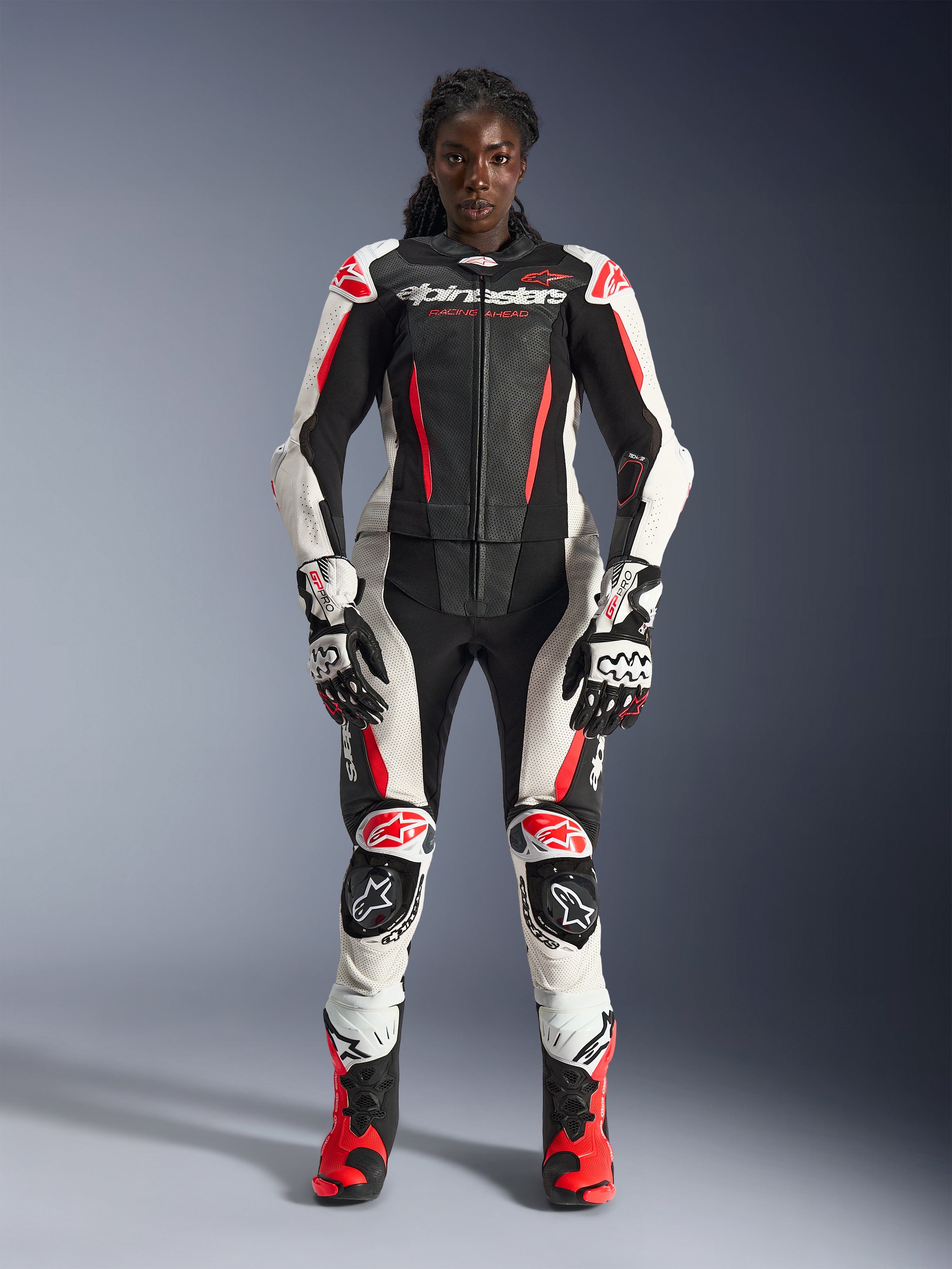Weibliches Model in Alpinestars Stella GP-R7 2-Teiler-Lederanzug in Schwarz, Weiß und Fluo-Rot, komplett mit passenden Handschuhen und Rennstiefeln, schützende Protektoren und perforierte Lederbahnen, professionelle Motorrad-Rennausrüstung