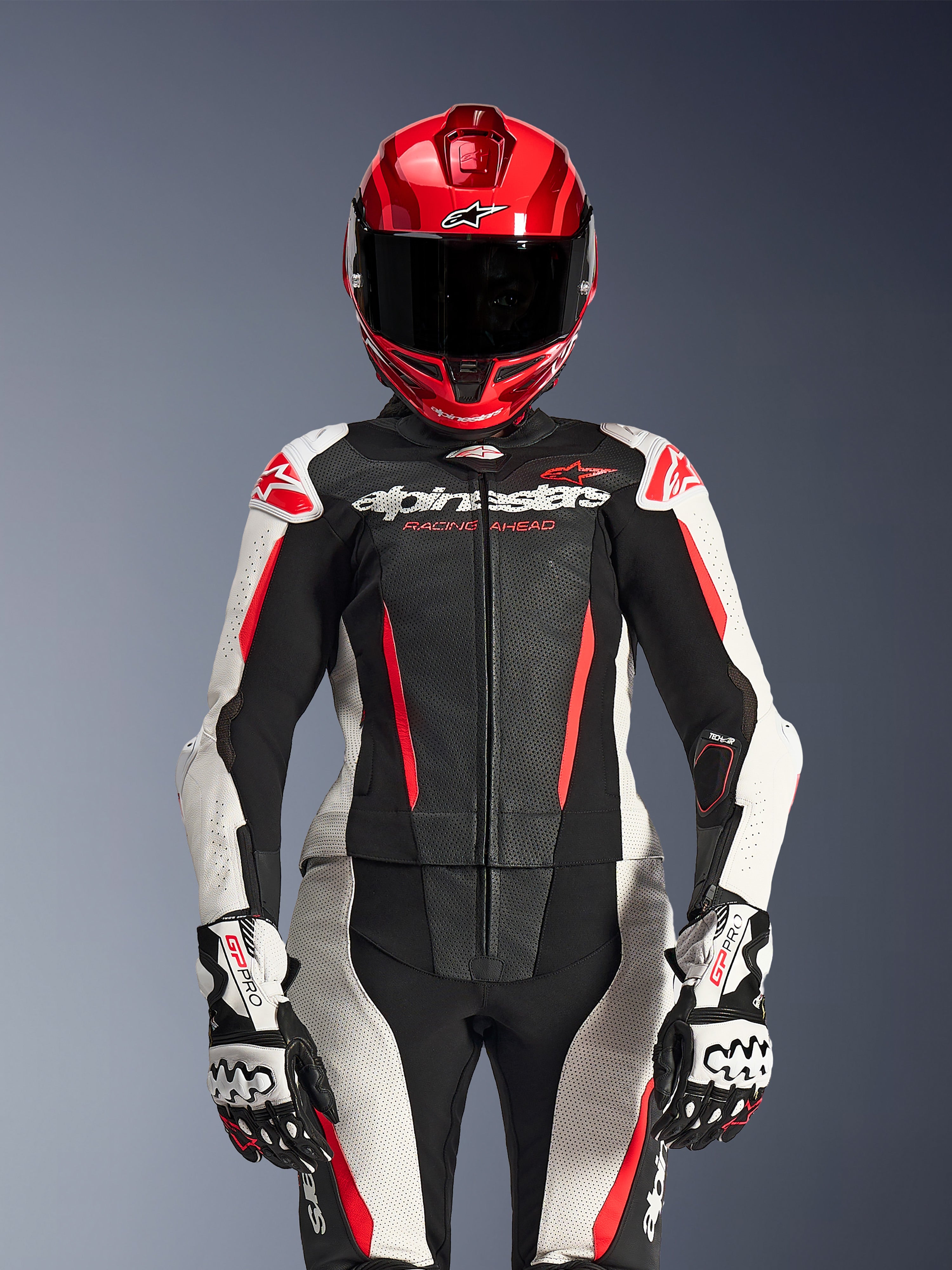 Alpinestars Stella GP-R7 2-Teiler-Lederanzug für Damen, Schwarz, Weiß und Fluo-Rot, mit perforierten Paneelen und 'Racing Ahead'-Branding, roter Helm, weiß-schwarze GP Pro Handschuhe, professionelle Rennsport-Ausrüstung