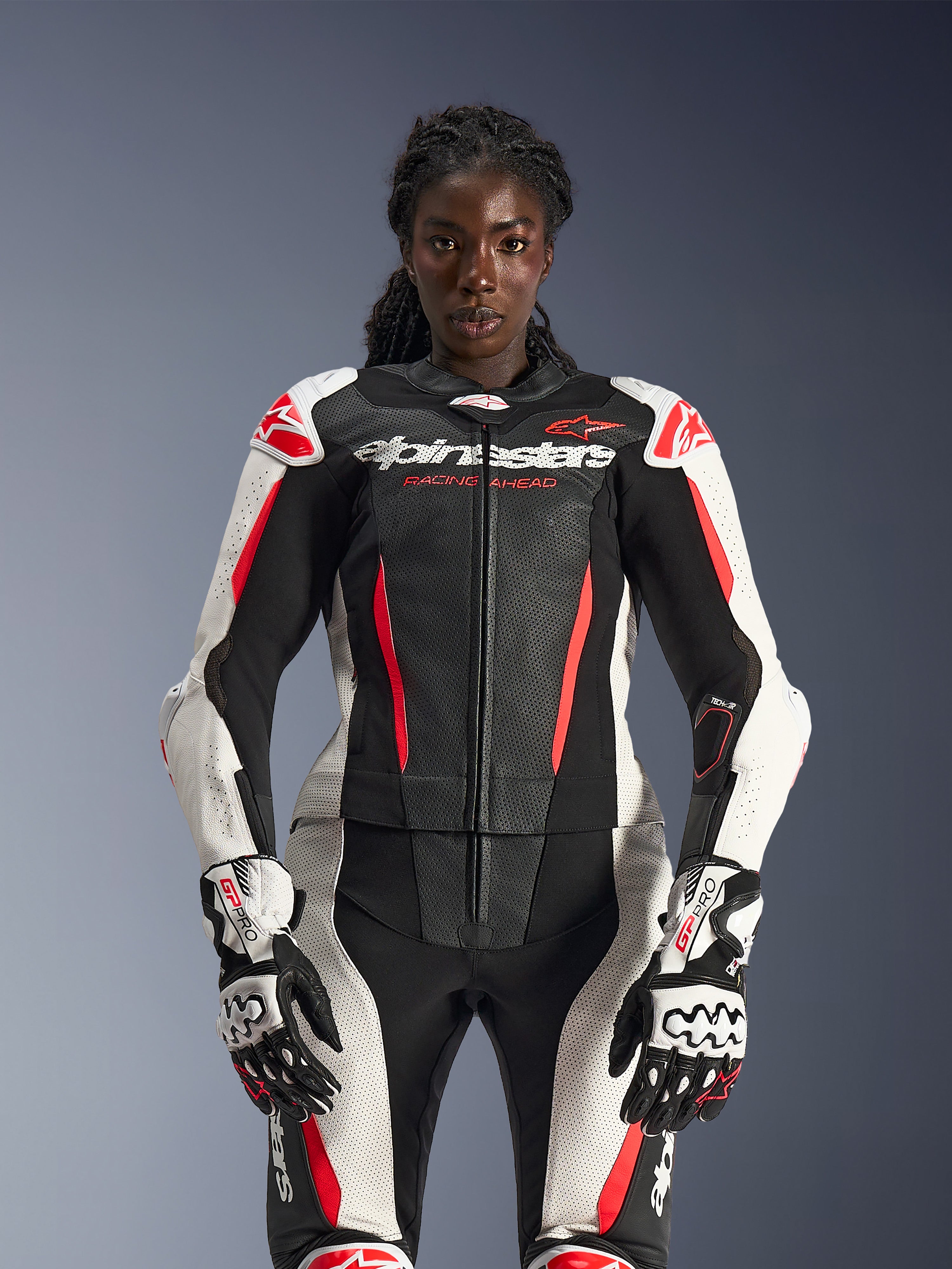 Weibliches Model im Alpinestars Stella GP-R7 2-Teiler-Lederanzug in Schwarz, Weiß und Red Fluo, mit perforierten Lederbahnen und passenden GP Pro Handschuhen, anatomisch optimiert für Fahrerinnen.
