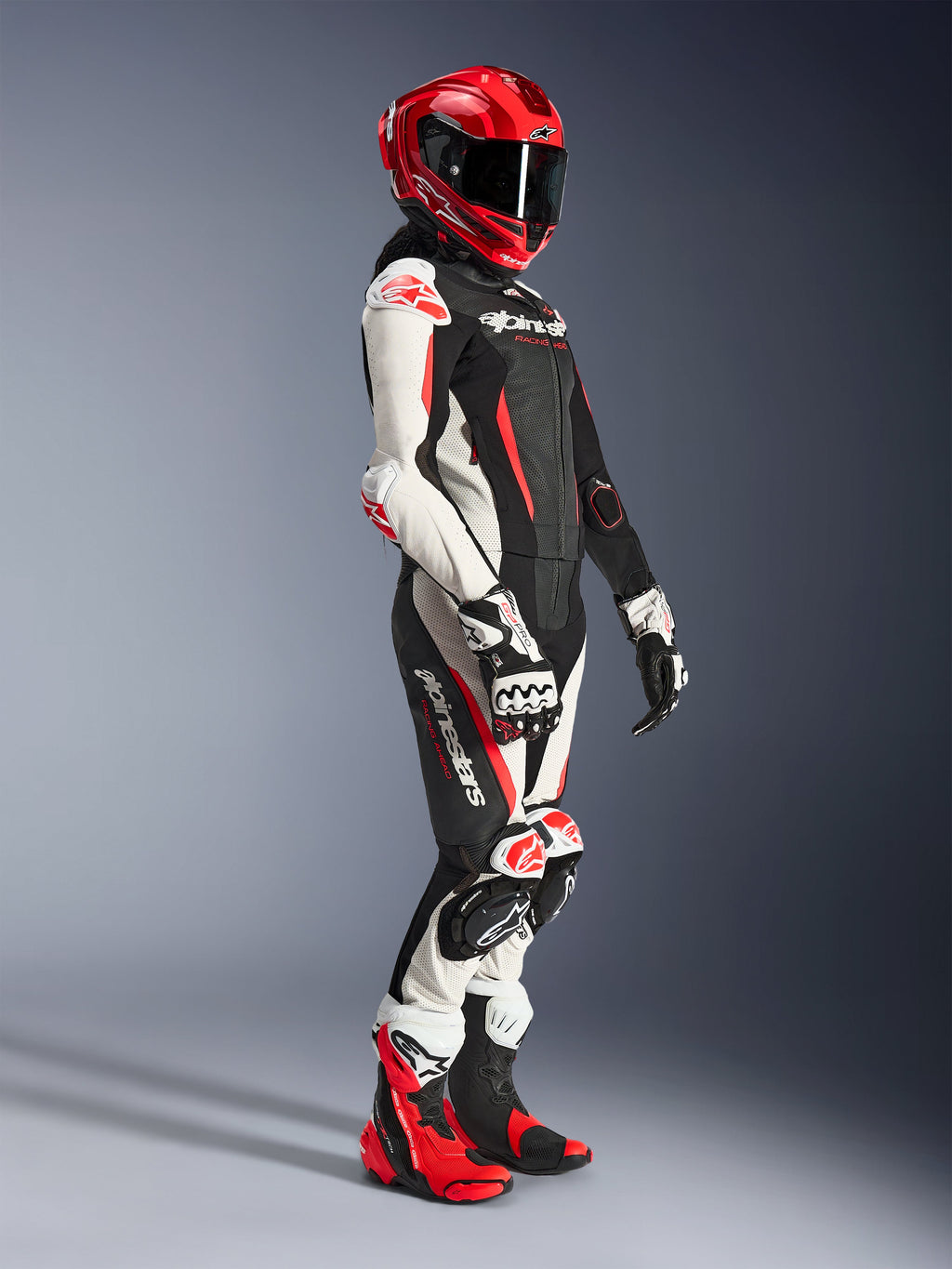 Motorradfahrerin im Alpinestars Stella GP-R7 2-Teiler-Lederanzug in Schwarz, Weiß und Fluo-Rot, kombiniert mit einem roten Integral-Rennhelm, passenden Handschuhen und Performance-Stiefeln, professionelle Motorrad-Rennausrüstung für Damen.