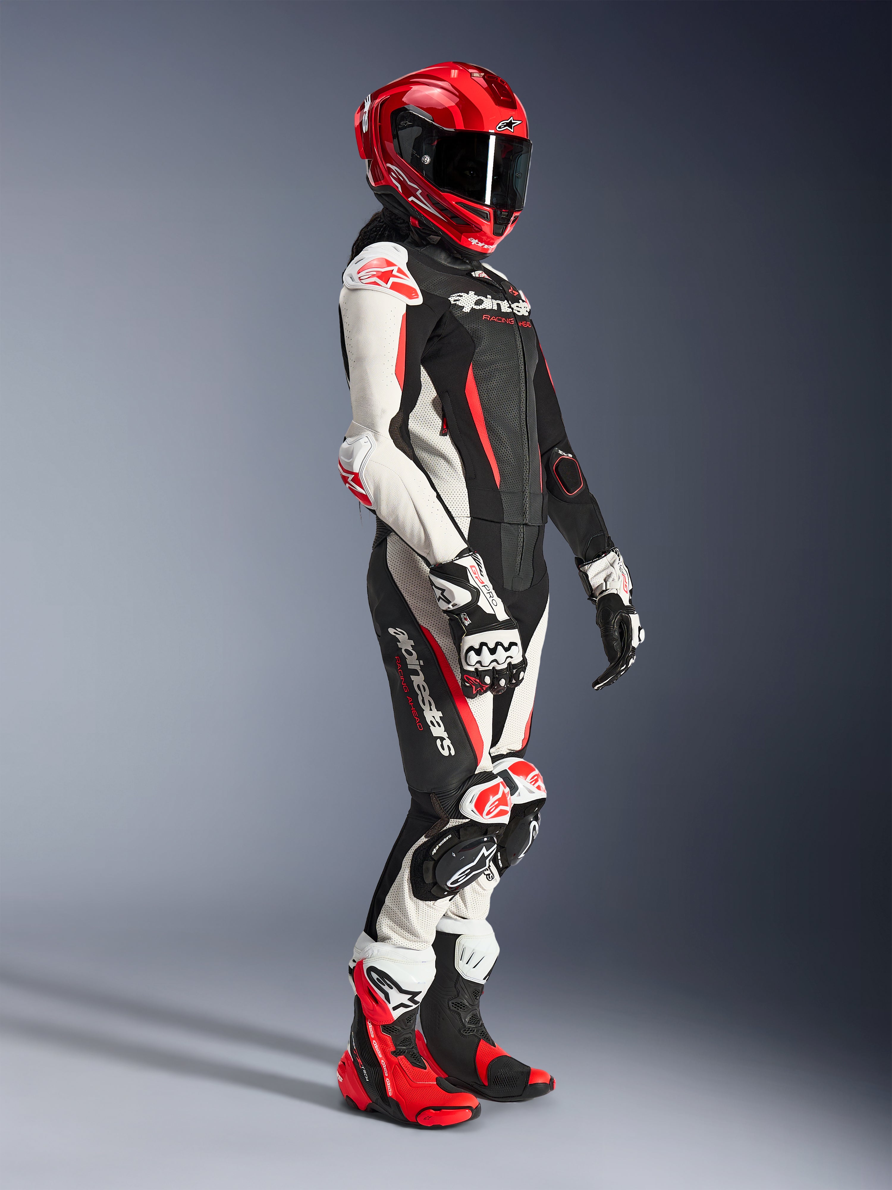Motorradfahrerin im Alpinestars Stella GP-R7 2-Teiler-Lederanzug in Schwarz, Weiß und Fluo-Rot, kombiniert mit einem roten Integral-Rennhelm, passenden Handschuhen und Performance-Stiefeln, professionelle Motorrad-Rennausrüstung für Damen.
