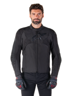 Alpinestars T-Jaws V4 Wasserdichte Jacke, schwarze Motorrad-Sportjacke, getragen von einem Mann mit schwarzen Handschuhen mit roten Akzenten, verstärkte Schultern und Arme, elegantes Design mit dezentem Brustlogo, textile Allwetter-Fahrerausrüstung