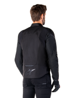 Alpinestars T-Jaws V4 Wasserdichte Jacke, Schwarz, Rückansicht am Modell, mit reflektierendem Alpinestars-Logo am unteren Rücken, verlängerte Rückenpartie für mehr Abdeckung, sportliches Textildesign mit farblich abgestimmten Einsätzen, getragen mit schwarzen Motorradhandschuhen