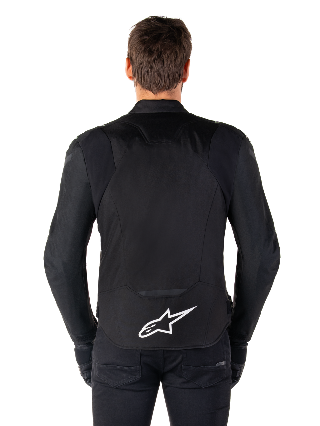 Alpinestars T-Jaws V4 Waterproof Jacke, schwarze Motorradjacke in der Rückansicht, mit weißem Alpinestars-Logo auf dem unteren Rücken, verlängerte Rückenpartie für optimale Abdeckung, getragen von Modell mit schwarzer Hose und Handschuhen, sportliches Textildesign