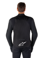 Alpinestars T-Jaws V4 Waterproof Jacke, schwarze Motorradjacke in der Rückansicht, mit weißem Alpinestars-Logo auf dem unteren Rücken, verlängerte Rückenpartie für optimale Abdeckung, getragen von Modell mit schwarzer Hose und Handschuhen, sportliches Textildesign