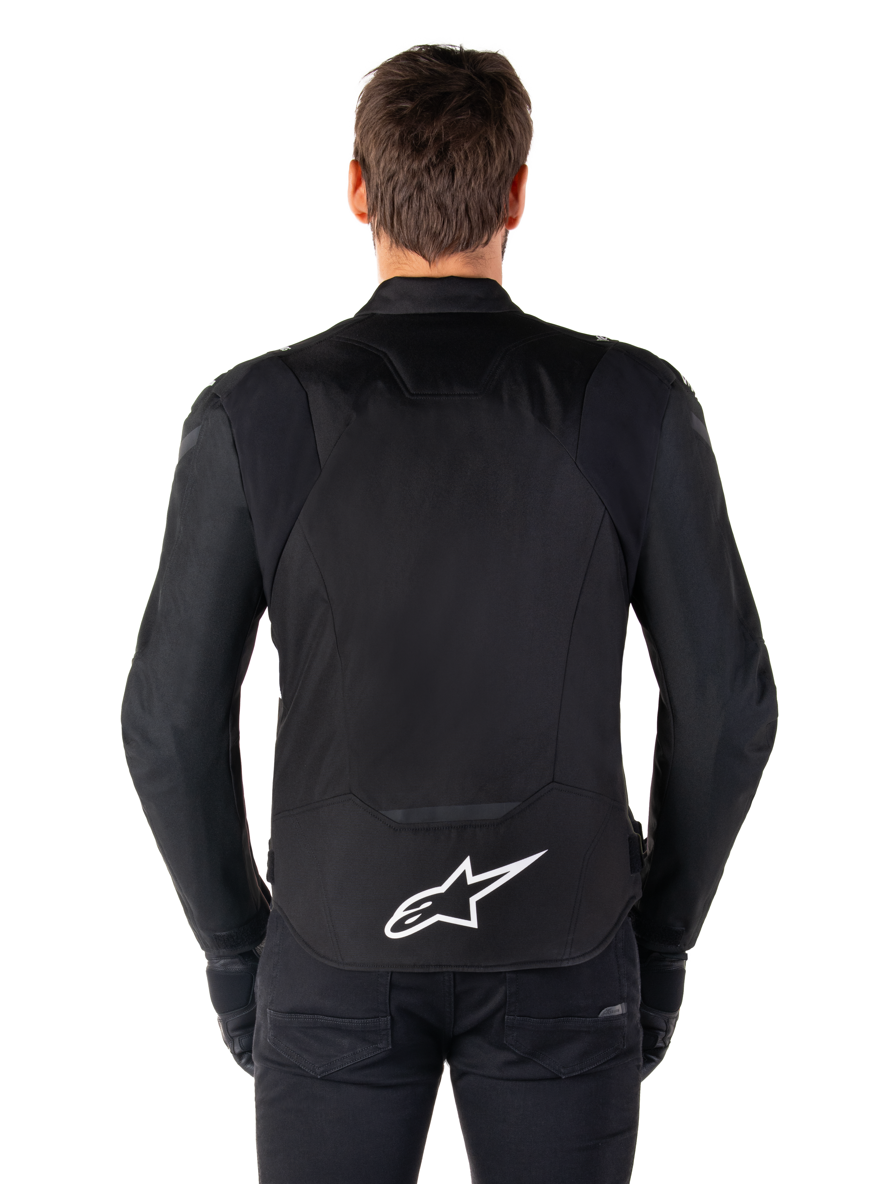Alpinestars T-Jaws V4 Waterproof Jacke, schwarze Motorradjacke in der Rückansicht, mit weißem Alpinestars-Logo auf dem unteren Rücken, verlängerte Rückenpartie für optimale Abdeckung, getragen von Modell mit schwarzer Hose und Handschuhen, sportliches Textildesign